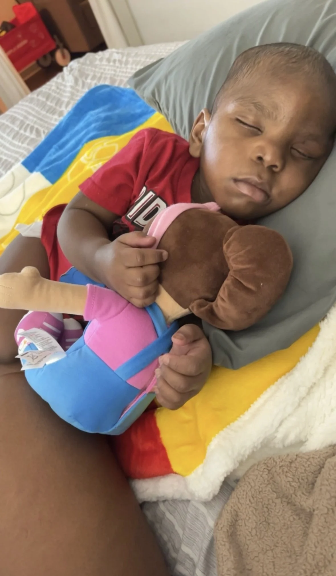A young boy with short hair sleeping on a bed, hugging a plush toy with brown, pink, and blue colors. The bed has a multicolored blanket with yellow, blue, and red sections.