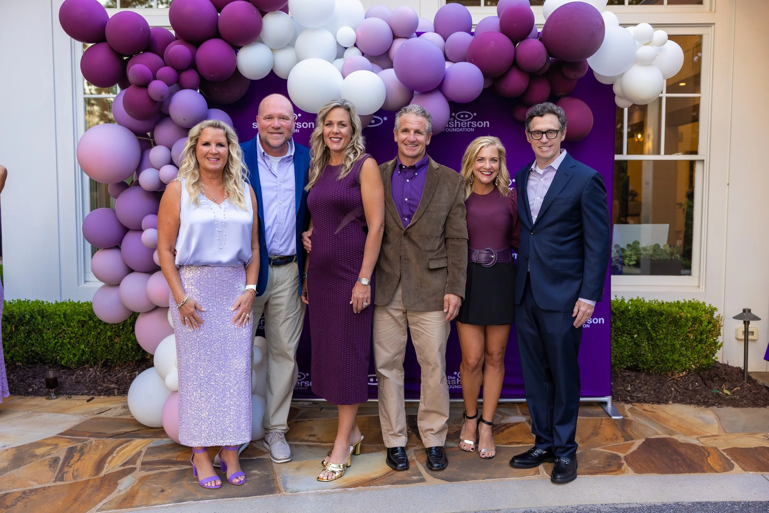 Group of six adults standing in front of a balloon arch with purple, pink, and white balloons, at an outdoor event. They are smiling and dressed in formal and semi-formal attire.