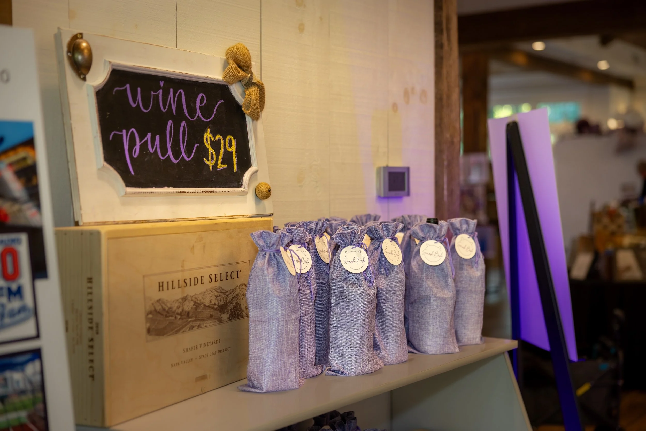 A display of small fabric gift bags with round tags, accompanied by a chalkboard sign that reads 'wine pull $29,' on a wooden shelf in a retail setting.