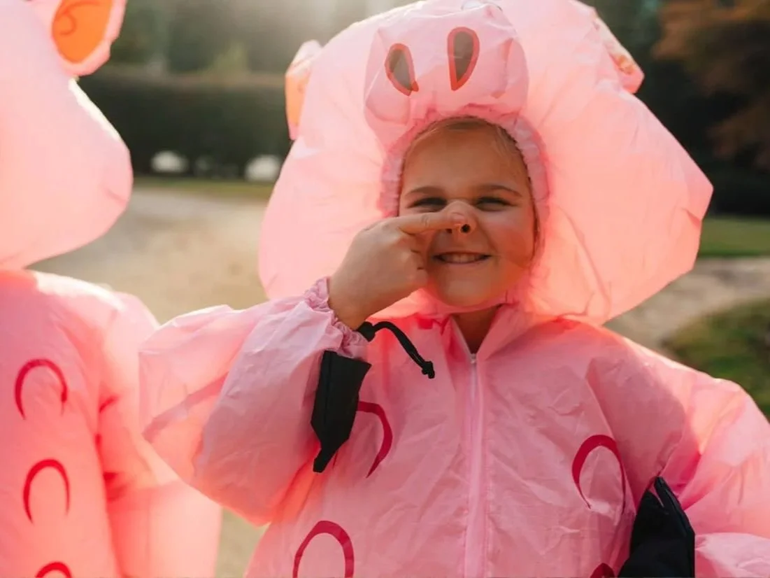 Child wearing a pink raincoat and hat with pig face design, smiling and holding nose with finger.