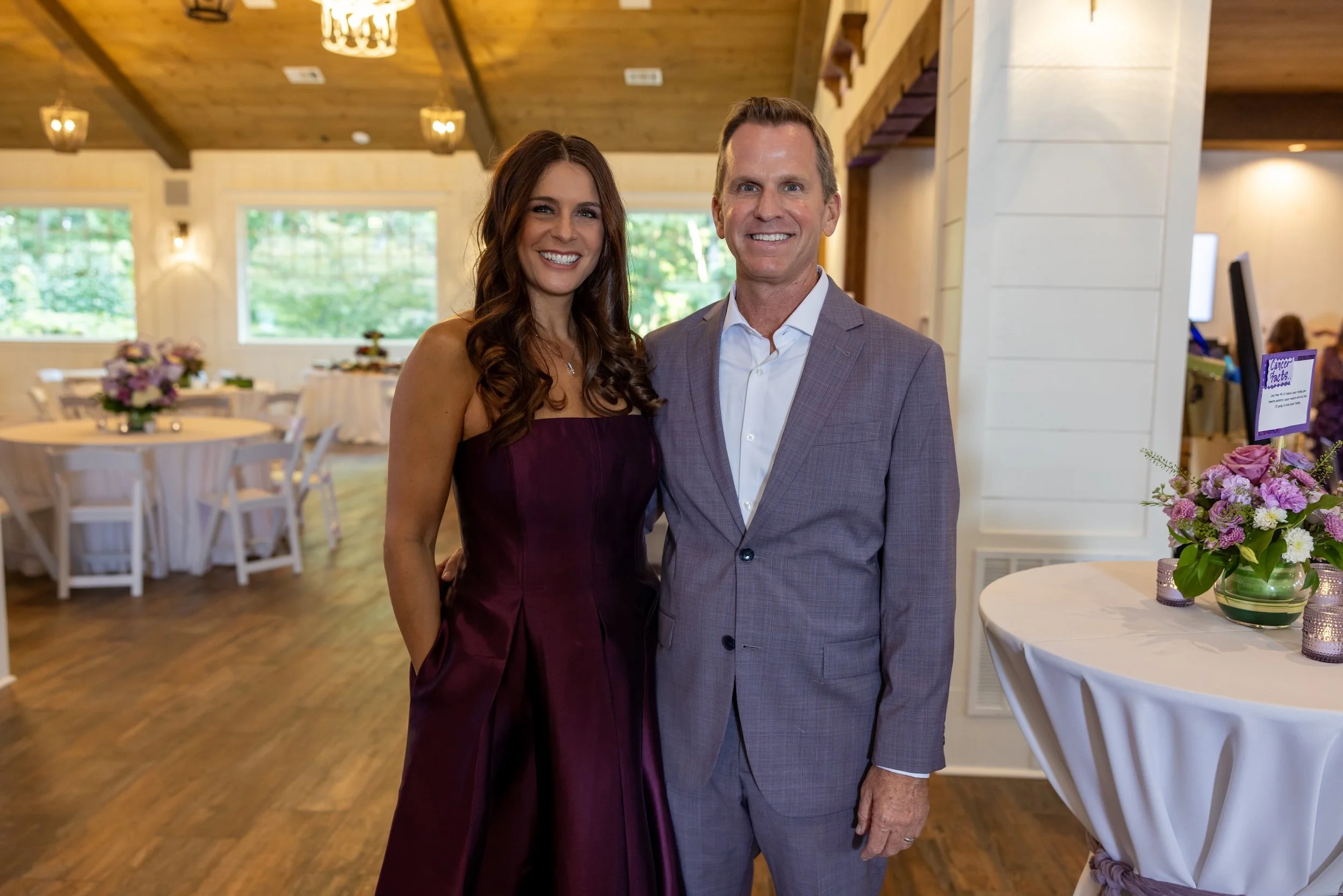 A smiling woman in a strapless purple dress and a smiling man in a gray suit standing in a decorated event space with tables, chairs, and floral arrangements.