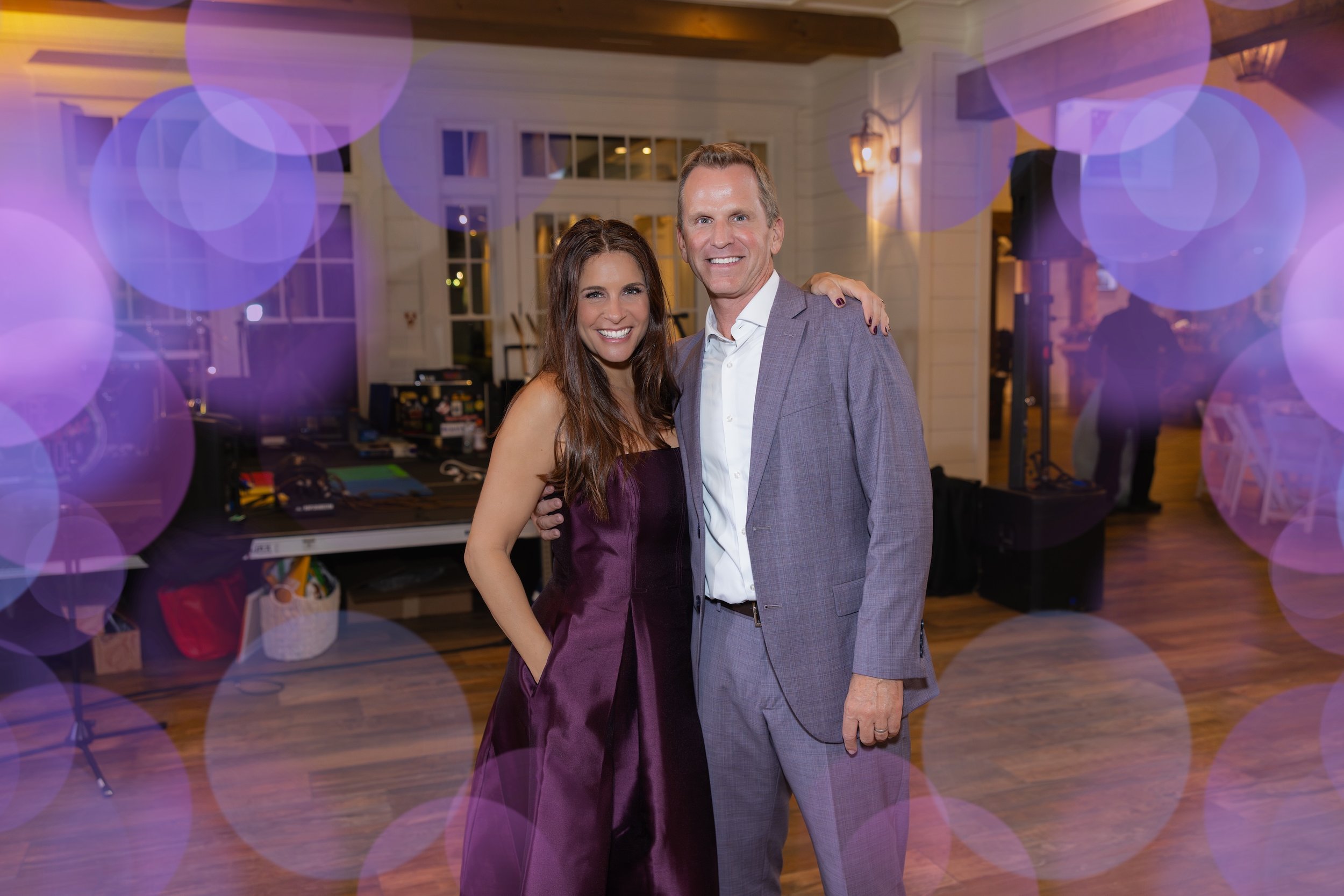 A man and woman smiling with their arms around each other at an indoor event, blurred purple bokeh circles overlay, background with windows and wooden ceiling beams.