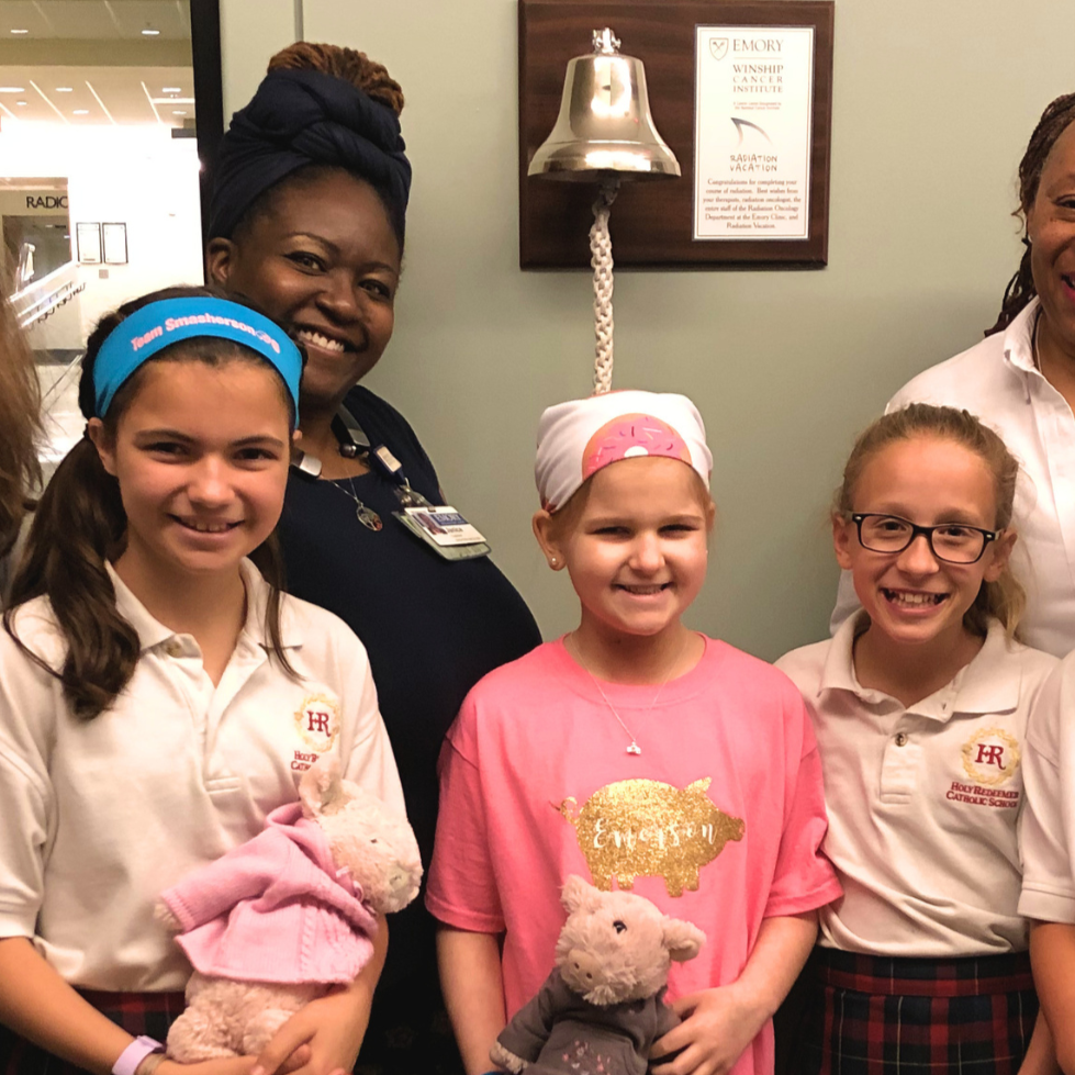 Group of four young girls and two women smiling for a photo indoors, with one girl holding a teddy bear and wearing a pink shirt with a golden pig graphic, in front of a green wall and a framed plaque.