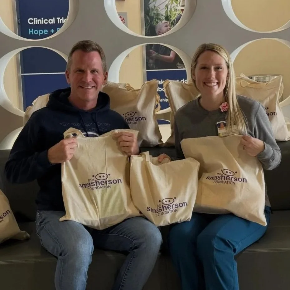 Two smiling individuals, a man and a woman, sitting on a bench surrounded by beige reusable bags with the logo of The Smשherson Foundation, in an indoor setting with circular wall decorations.