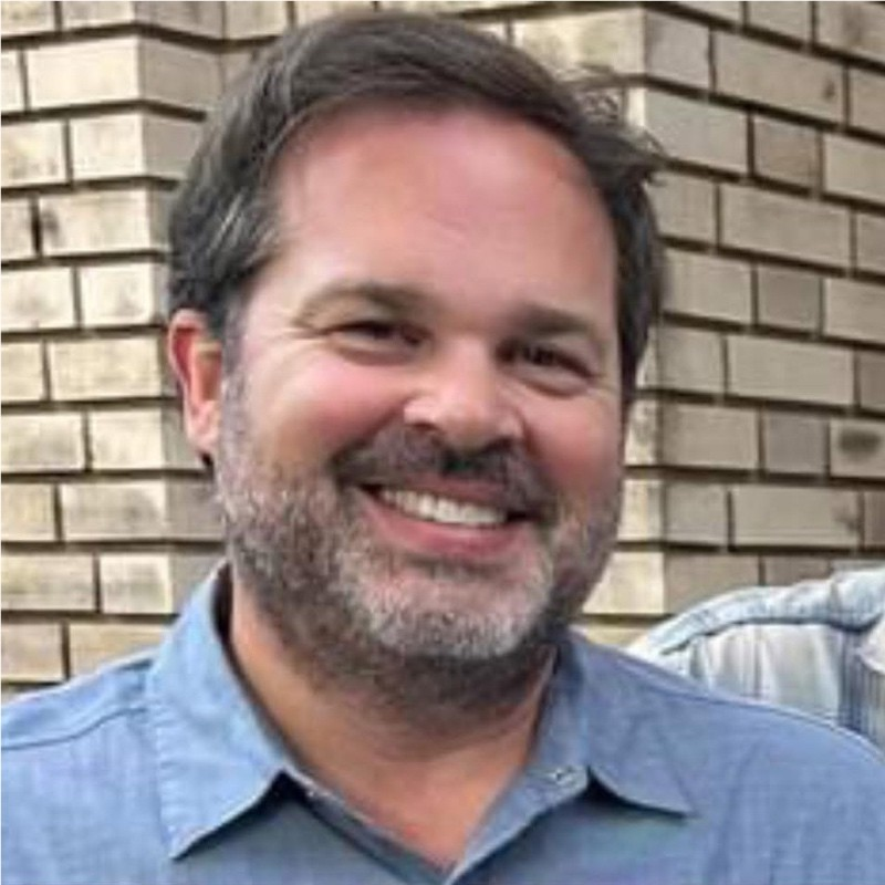 A man with dark hair, a beard, and a mustache smiling outdoors in front of a brick wall, wearing a blue shirt.