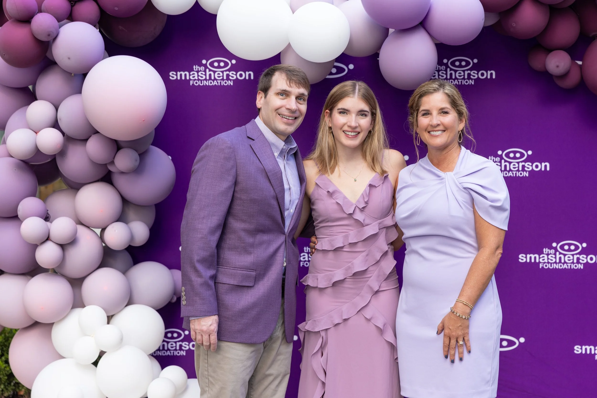 Three people standing in front of a purple backdrop with balloons, smiling at a foundation event. The backdrop has the logo 'the smasheron FOUNDATION' repeated multiple times.