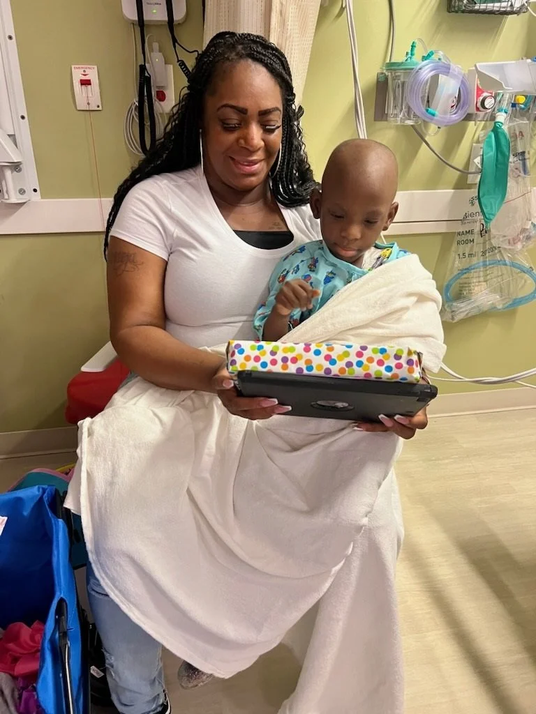 A woman and a young boy in a hospital room looking at a tablet. The woman is sitting in a hospital bed holding the boy, who is wrapped in a hospital blanket and wearing a hospital gown. Hospital equipment is visible in the background.