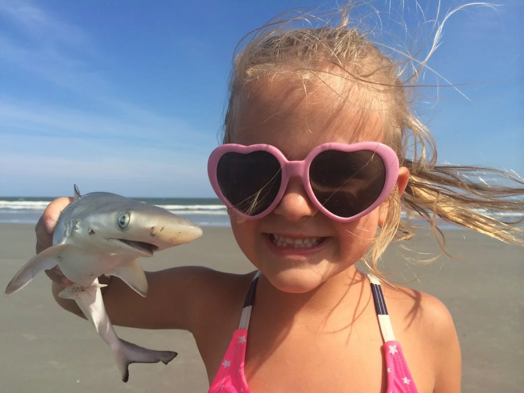 A happy young girl wearing pink heart-shaped sunglasses and a pink swimsuit, holding a small shark on a beach with the ocean and blue sky in the background.