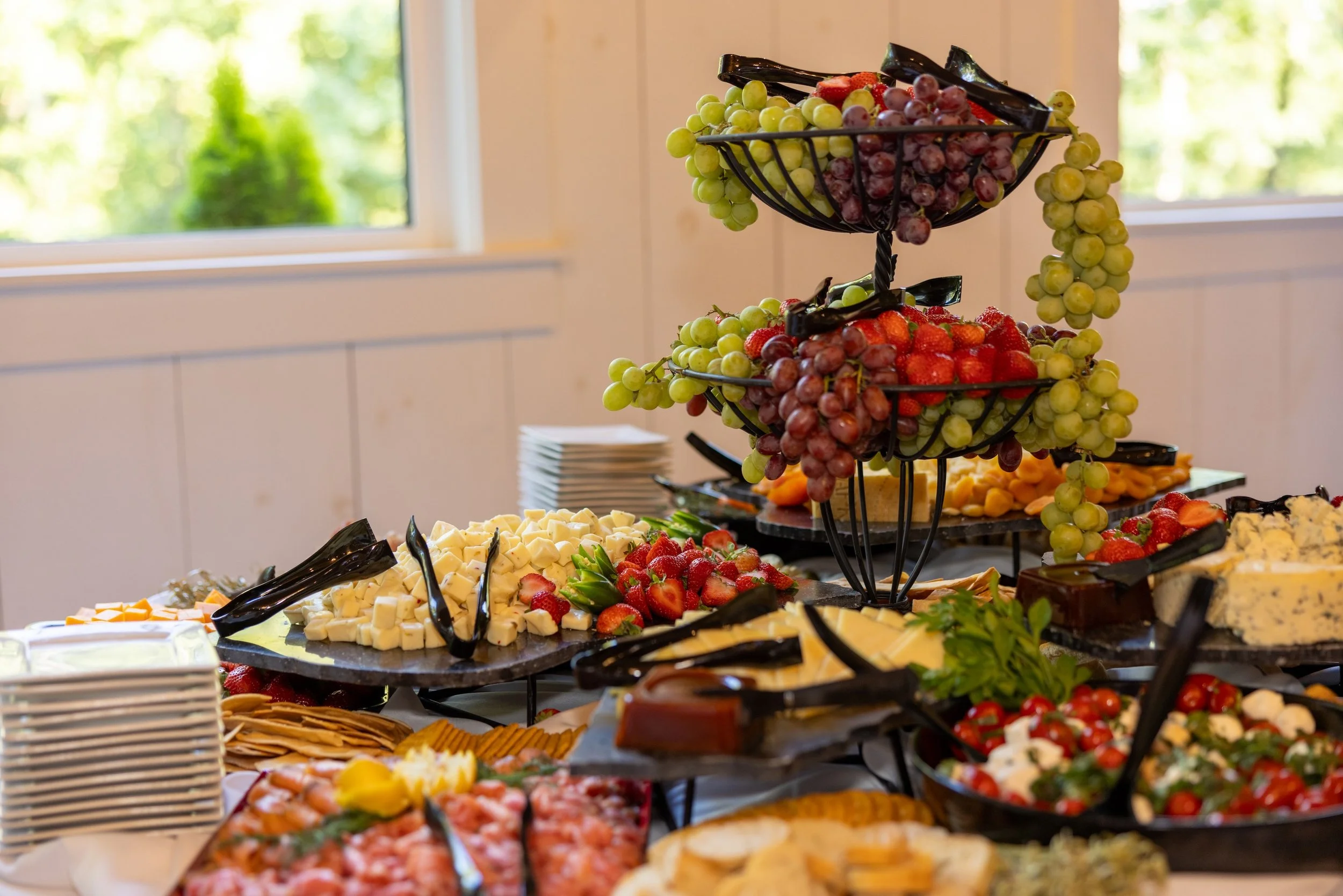 A multi-tiered display of various fresh fruits and cheeses, including strawberries, grapes, and sliced cheese on a buffet table, with windows showing greenery in the background.