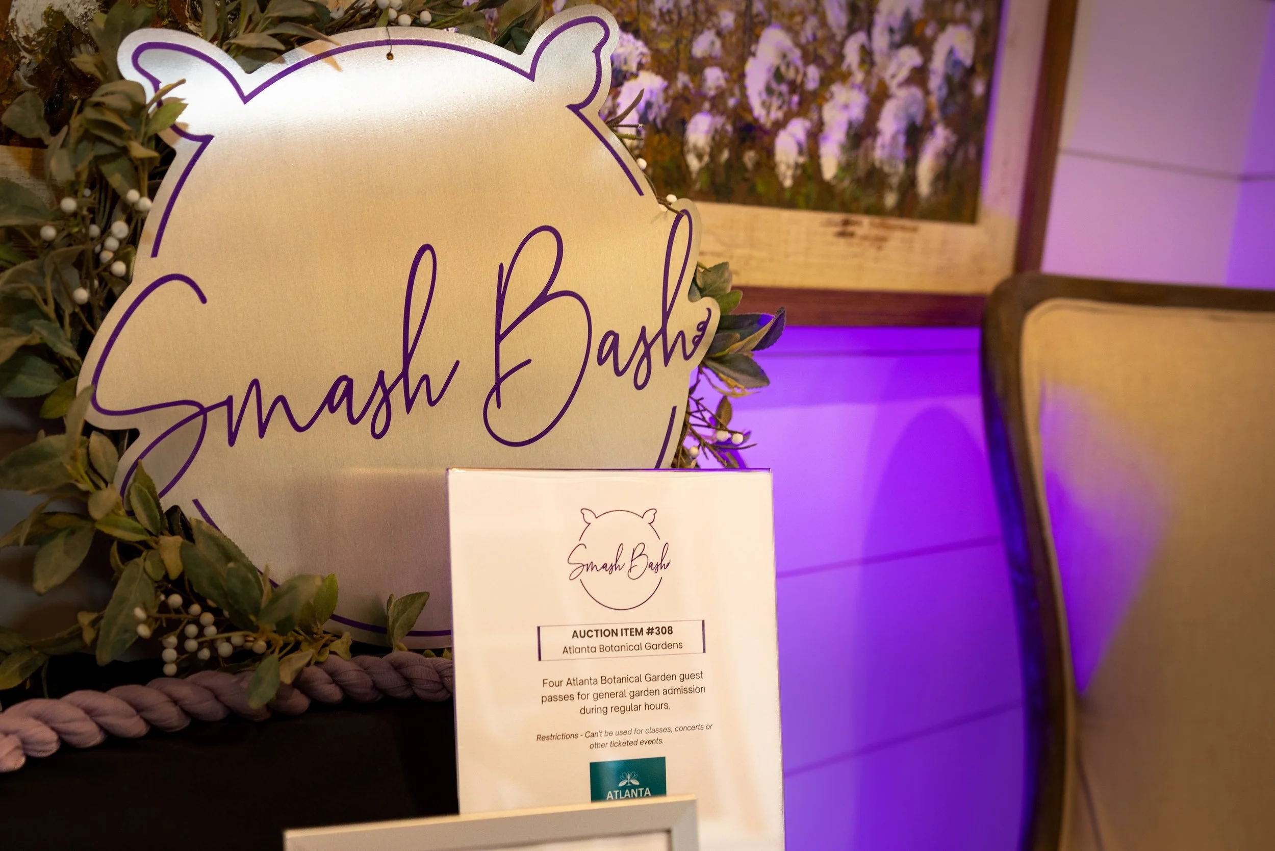 A decorative sign with the words "Splash Bash" written in purple cursive, surrounded by green foliage with small white decorative elements. In front of the sign, there is an informational card about an auction item from Atlanta Botanical Gardens, wit