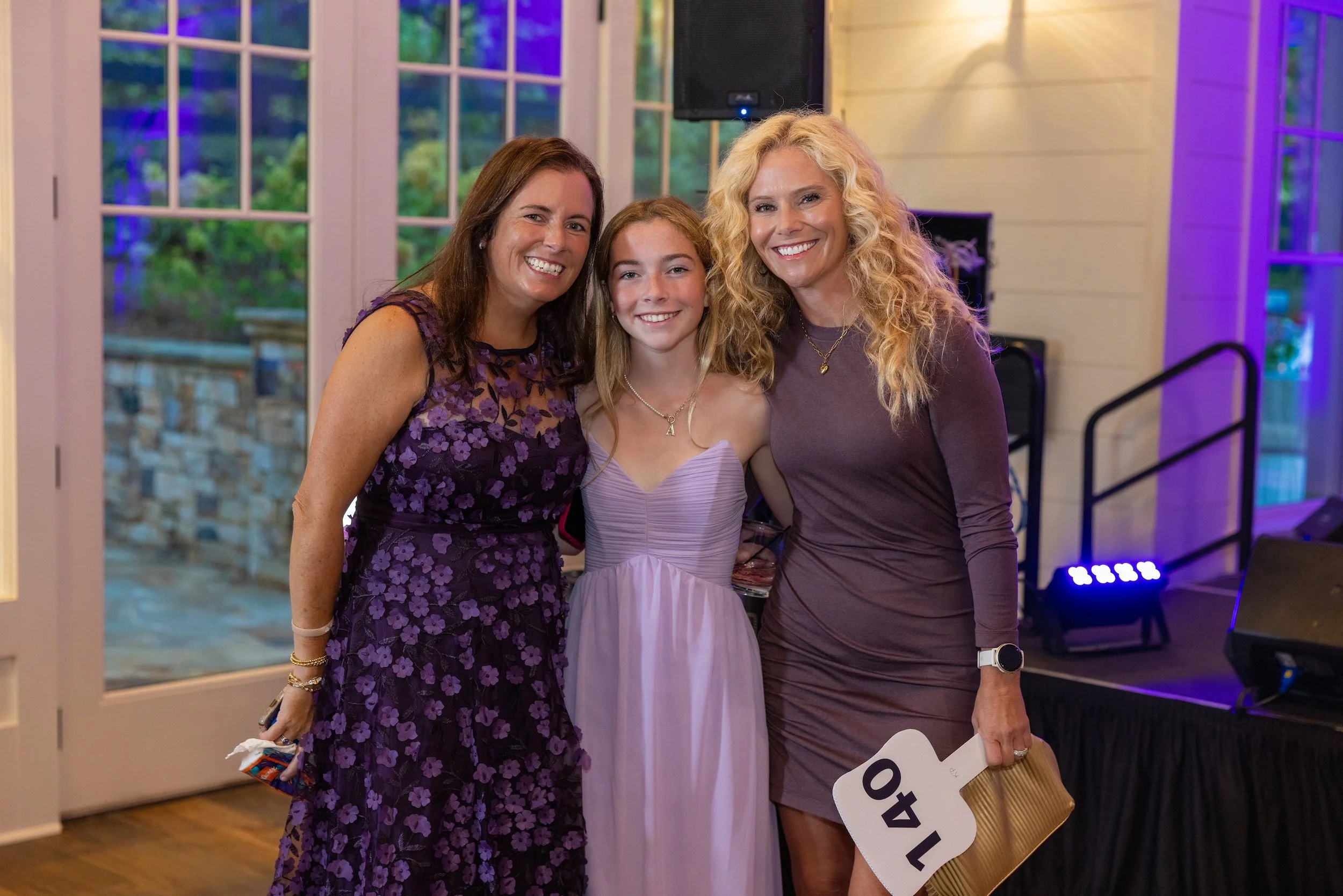 Three women and a girl smiling and posing together at a party or event, with semi-open windows showing greenery outside in the background.
