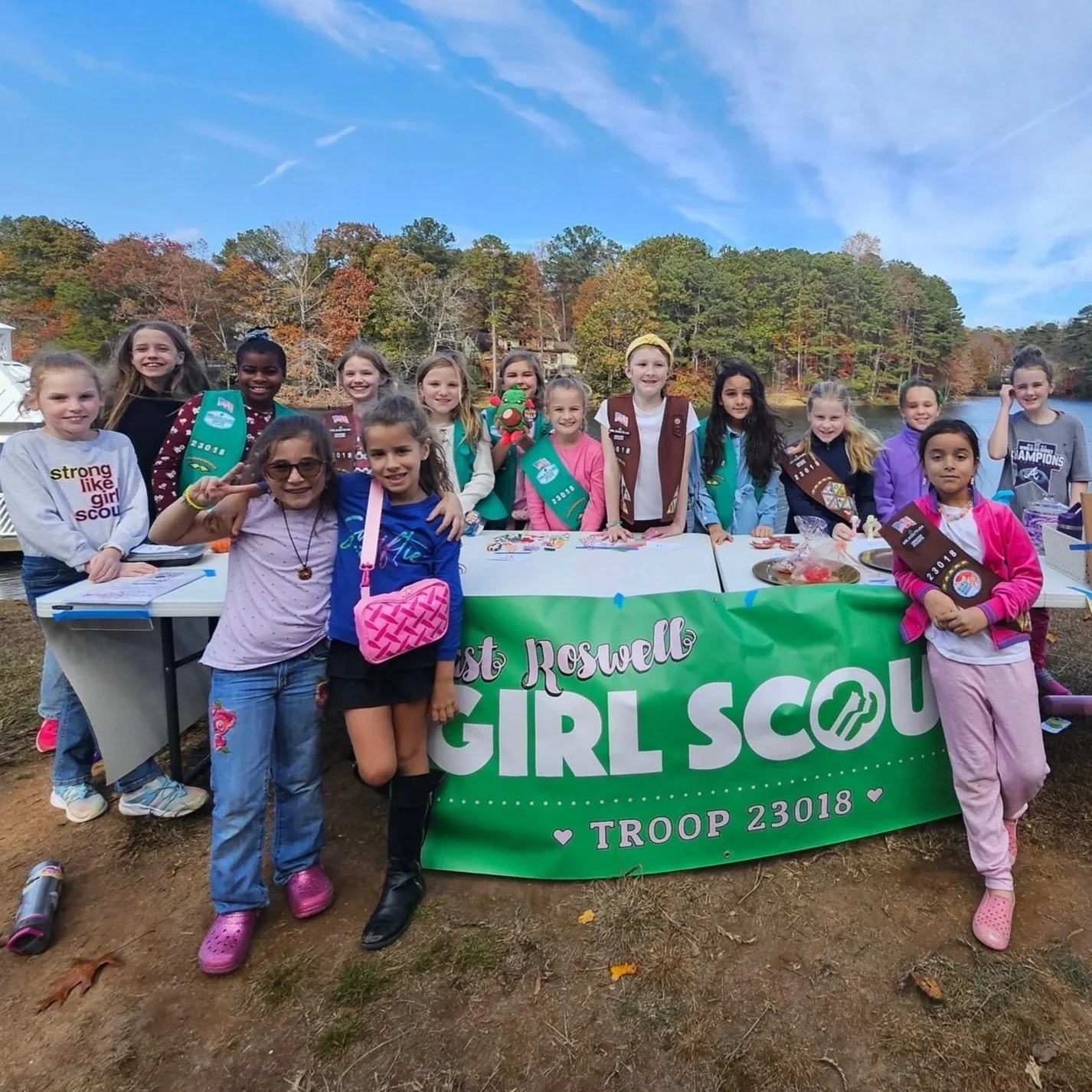 Group of young Girl Scouts smiling outdoors behind a table with a green "Girl Scout Troop 23018" banner, trees with fall foliage, and a lake in the background.