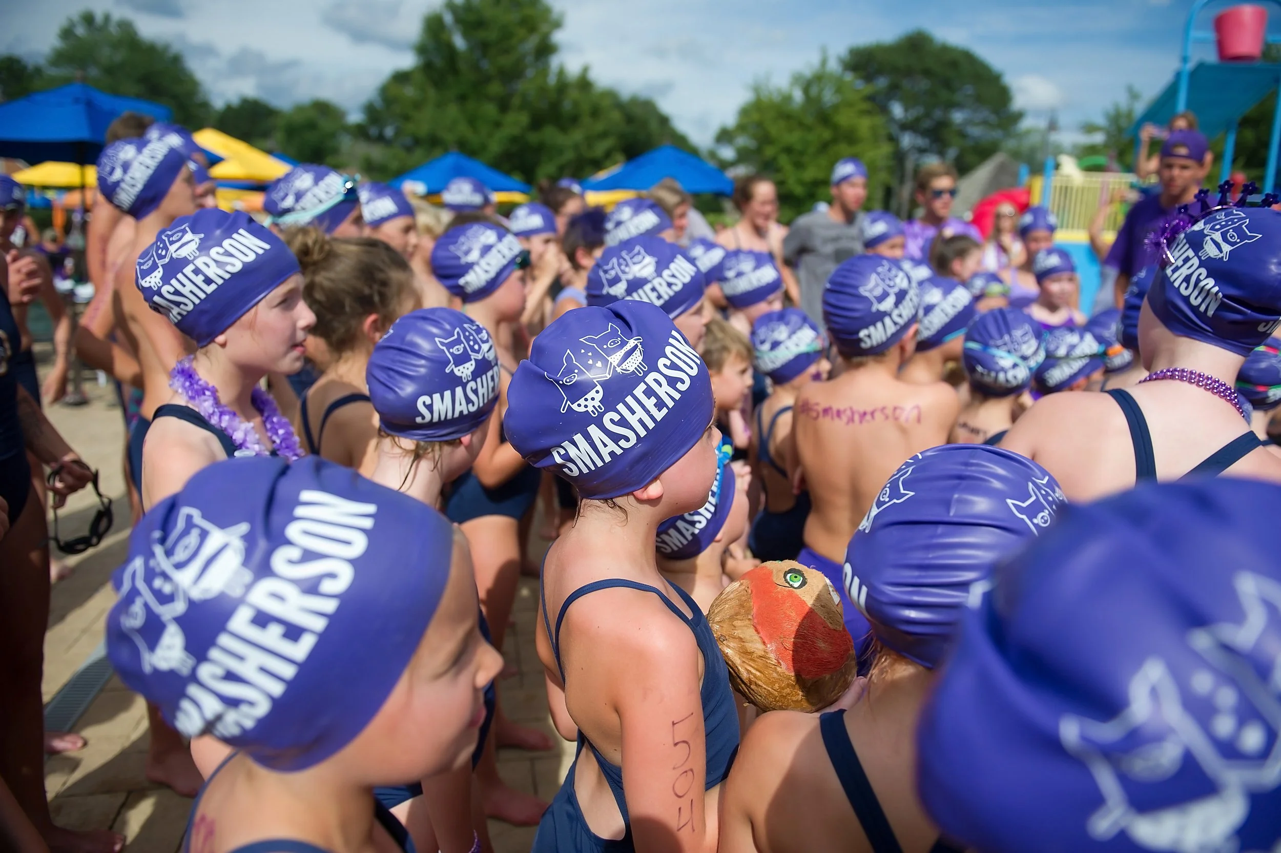A group of children wearing blue swim caps with the word 'SMASHERSON' printed on them, gathered outdoors under a partly cloudy sky, possibly preparing for a swimming competition or event.