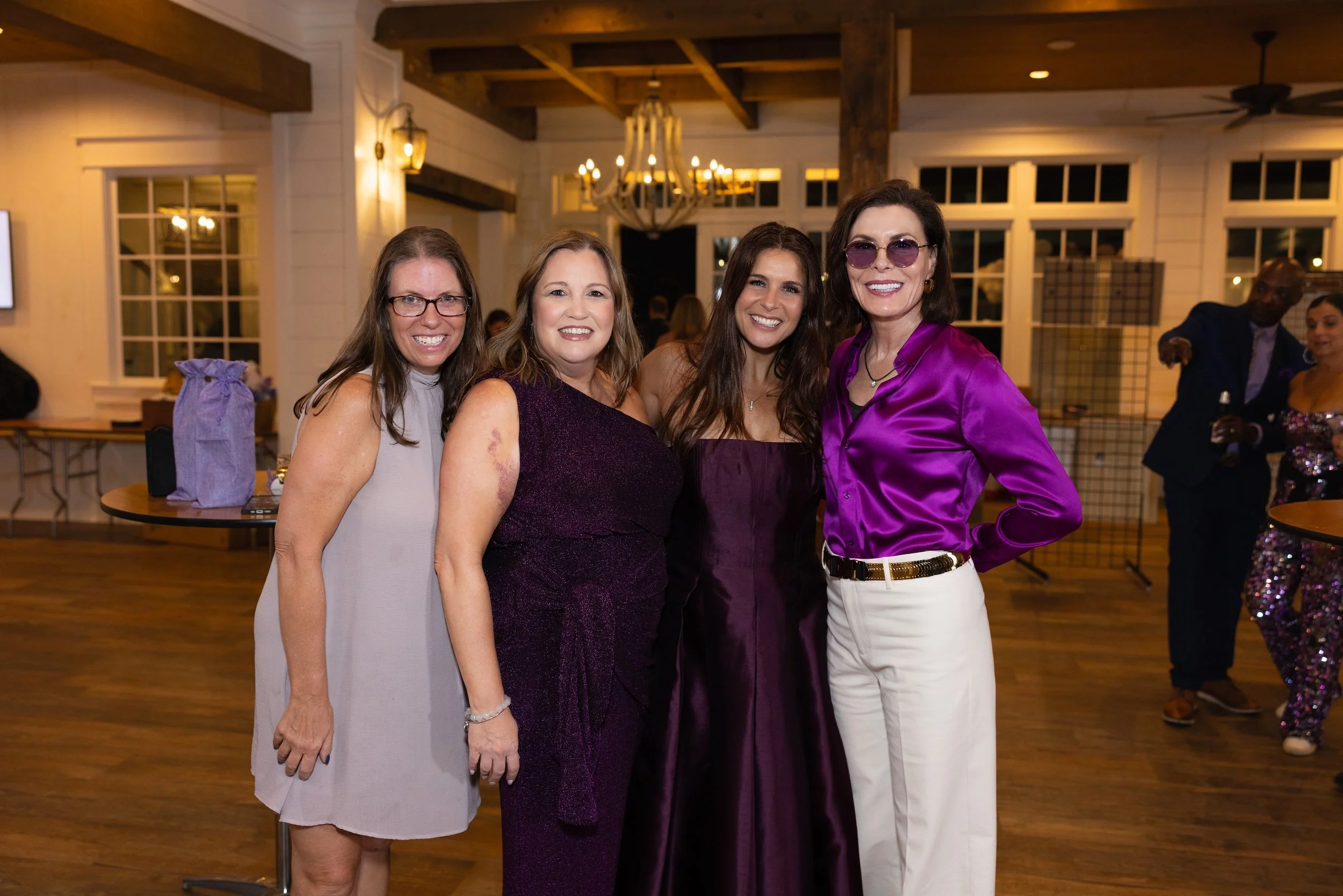 Four women posing together at a social event in a warmly lit hall with chandeliers and large windows.