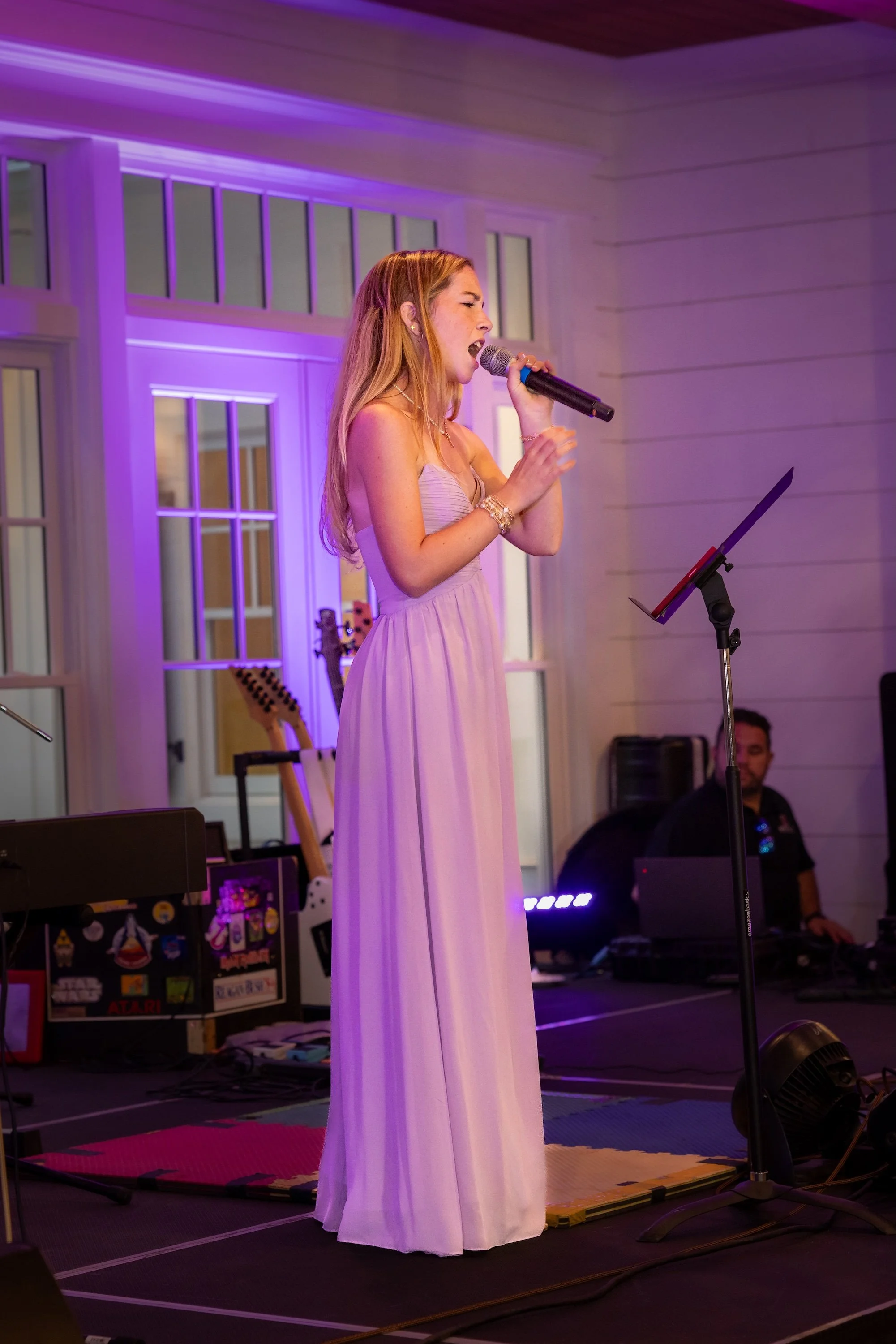 A woman in a strapless purple gown singing into a microphone on stage during a performance with purple lighting. There are guitars and music equipment in the background.