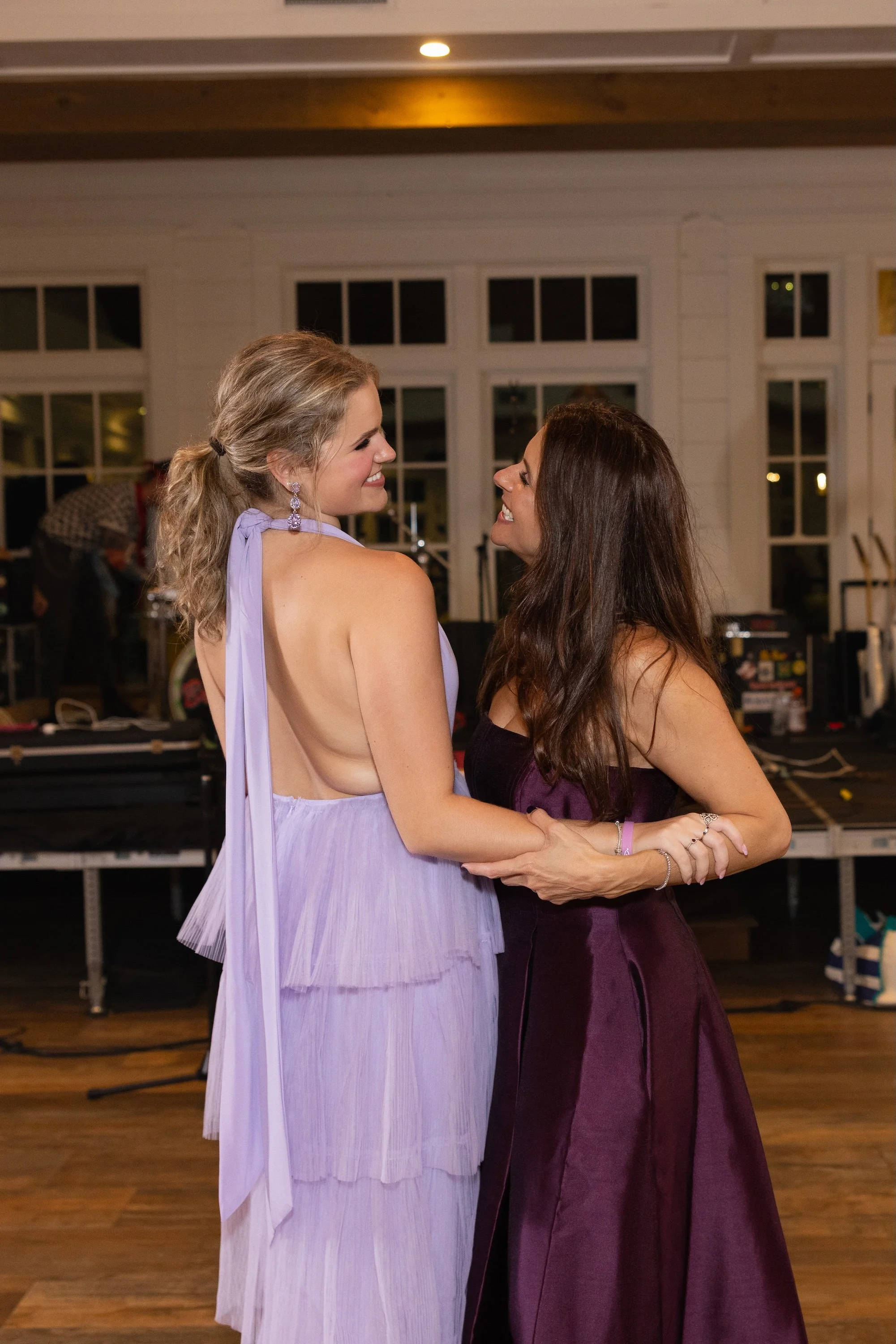 Two women in formal dresses holding hands and smiling at each other in an indoor event space.