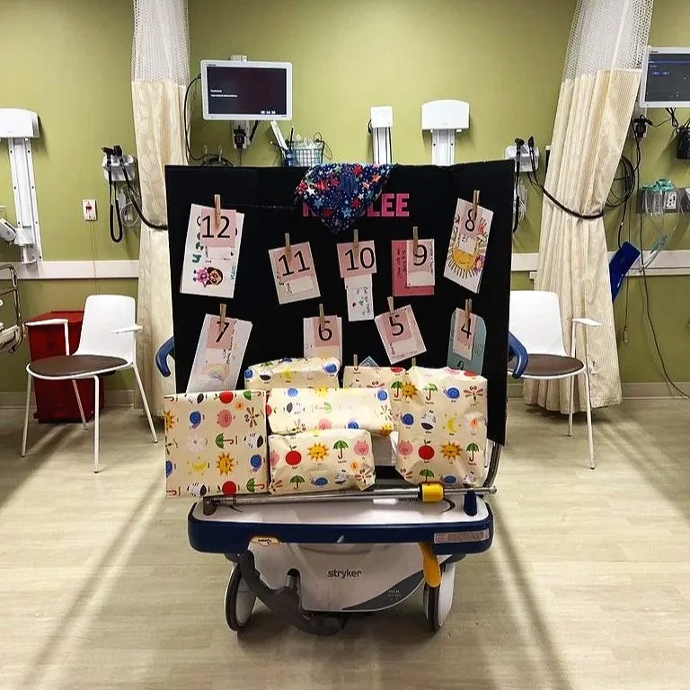 Hospital setting with a decorated advent calendar made of wrapped gifts on a wheeled hospital bed, numbered cards from 1 to 12 hanging on a black board, medical equipment on the walls, and chairs near curtains.
