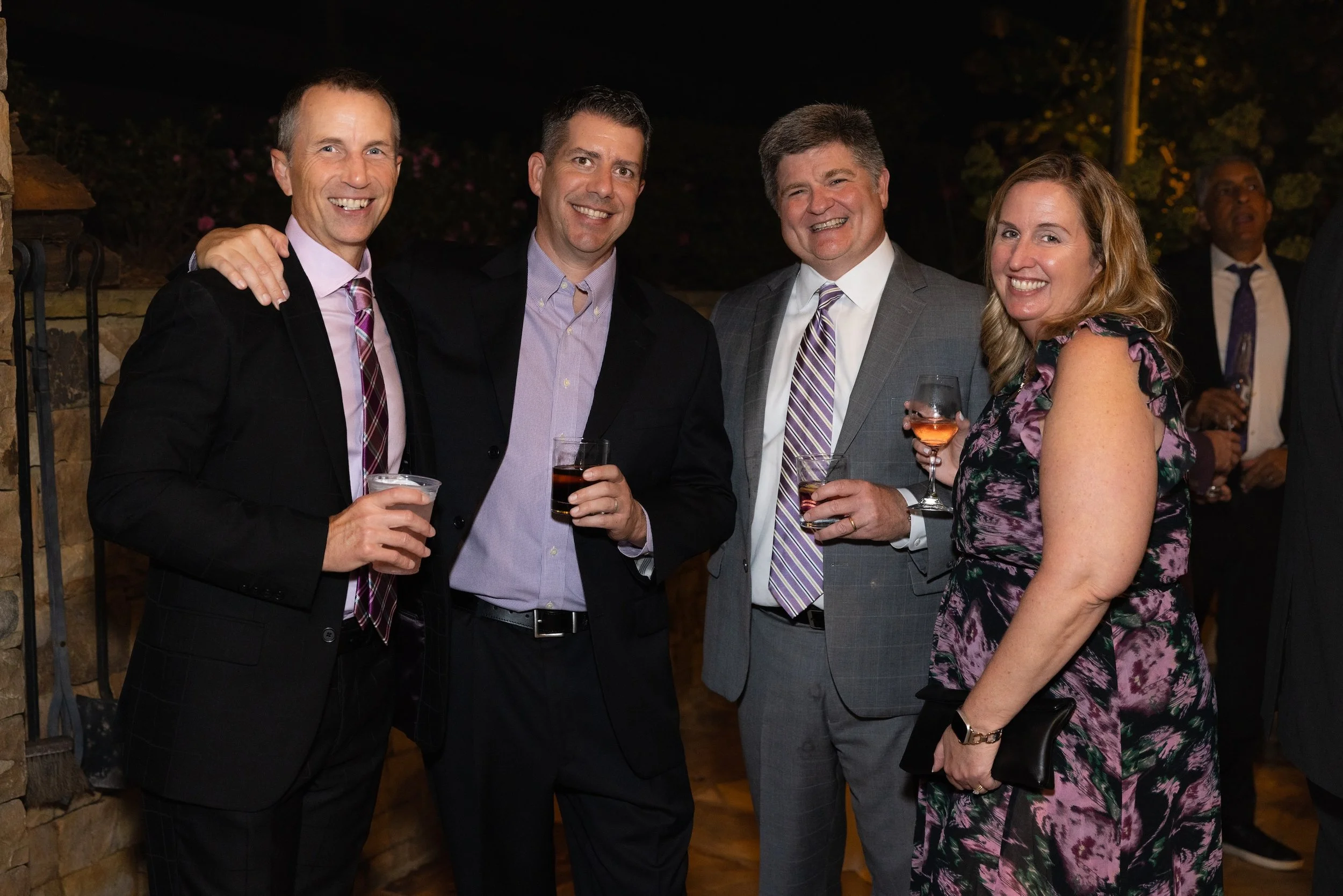 Four people, two men in suits, and a woman in a floral dress, smiling and holding drinks at a social gathering at night.
