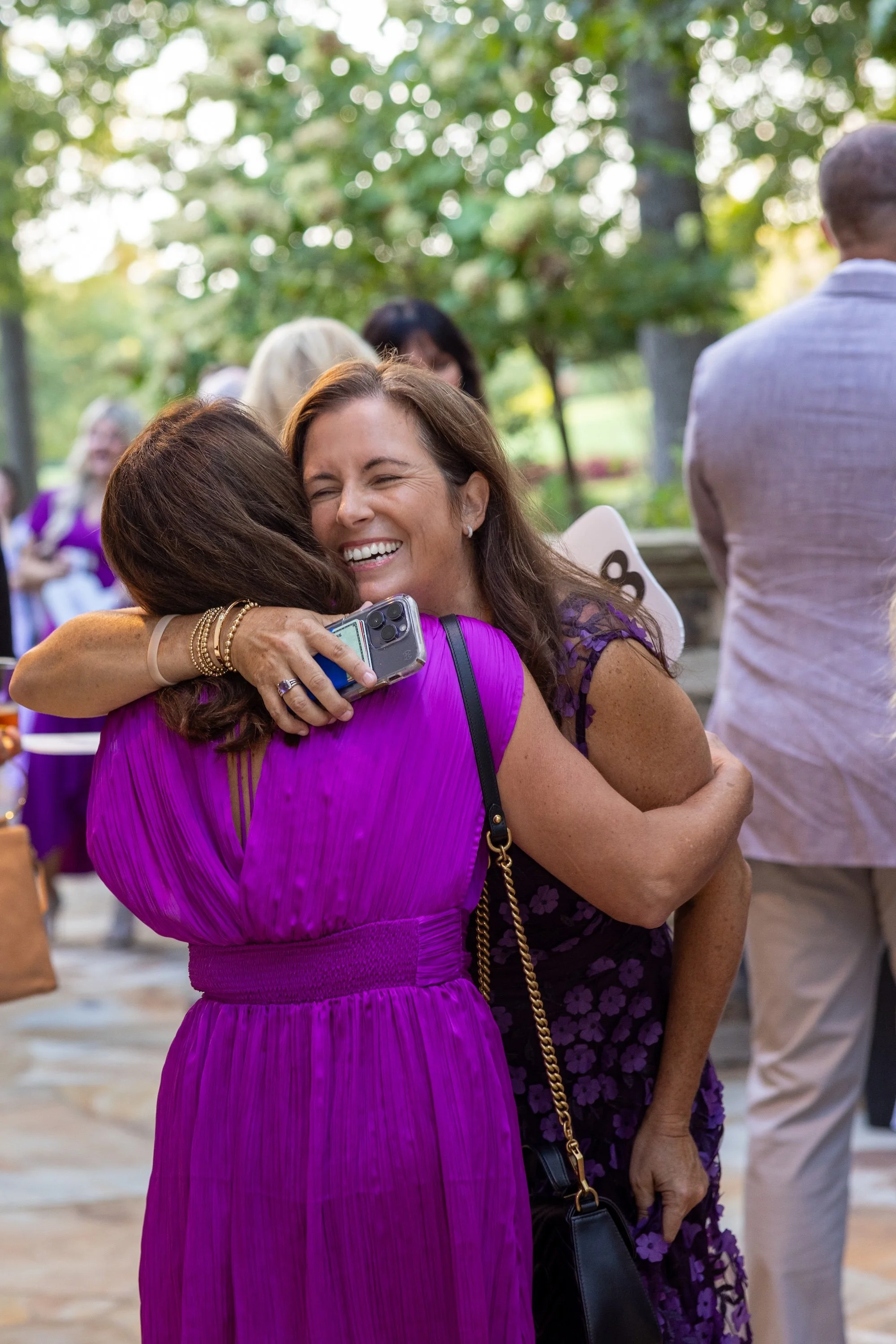 Two women hugging and smiling at an outdoor event; one wearing a bright purple dress, the other in a purple floral dress; other people are in the background.