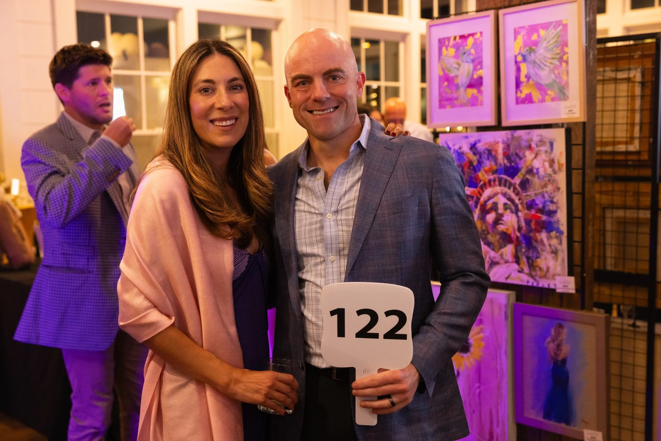 A smiling woman and man standing together at an art gallery event. The woman is wearing a light pink blazer and the man is in a gray plaid suit holding a sign numbered 122. In the background, a man in a checkered blazer is eating, and colorful artwor