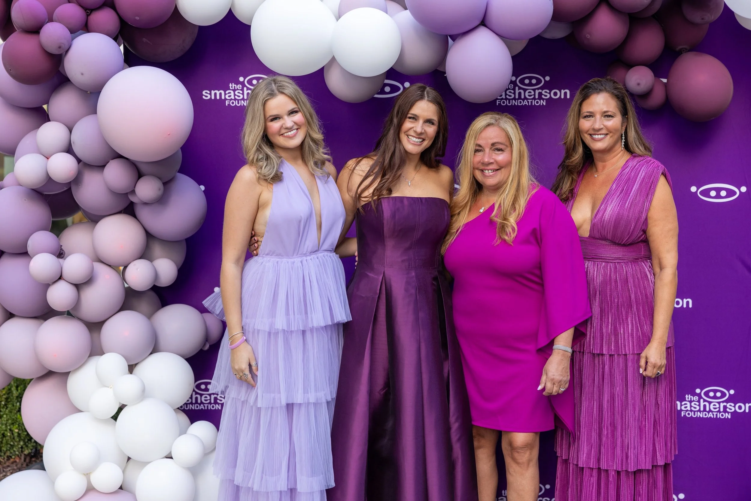 Four women in purple and pink dresses standing together at a charity event with balloon decorations and a purple backdrop that reads 'the Splasherson Foundation'.