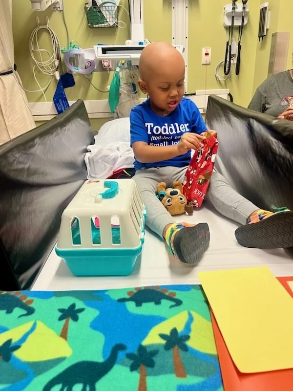 A young child with a bald head sitting on a hospital bed, looking at a red bag. The child is wearing a blue shirt that says "Toddler" and gray pants. There are toys near the child and medical equipment on the wall behind, indicating a hospital or clinic setting.