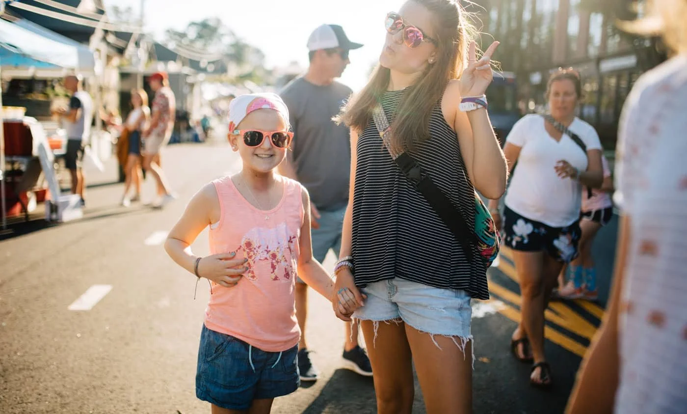 Two smiling girls, one younger and one older, holding hands at an outdoor fair or market during daytime. They are wearing sunglasses and casual summer clothing, with tents and other people in the background.