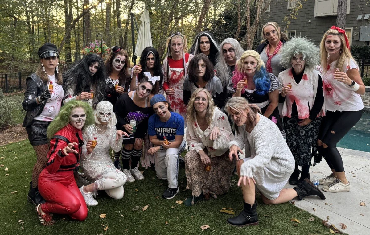 Group of women and children dressed in Halloween costumes, some with spooky makeup, posing outdoors in a backyard.