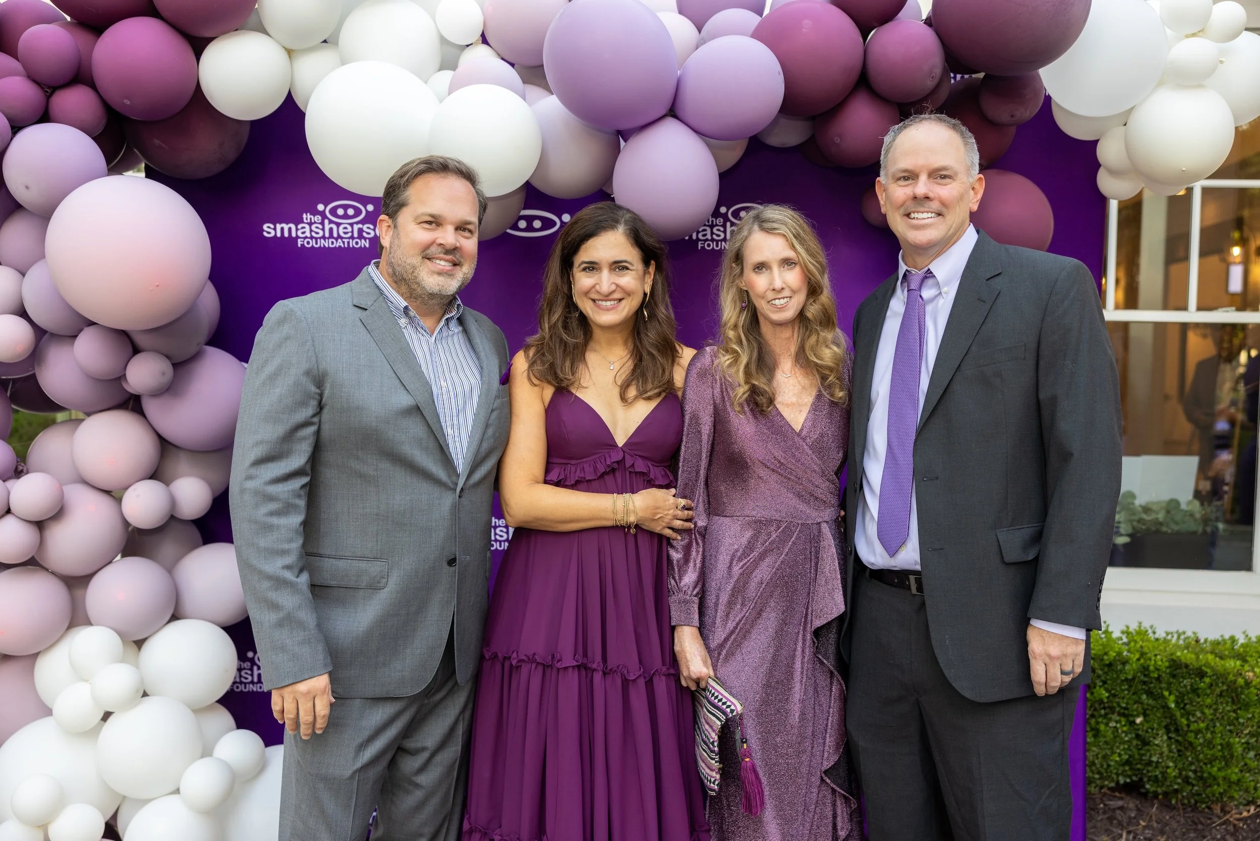 Four people standing in front of a purple backdrop decorated with pink, purple, and white balloons, with the logo of The Smasher Foundation visible behind them. They are dressed formally, smiling at the camera.