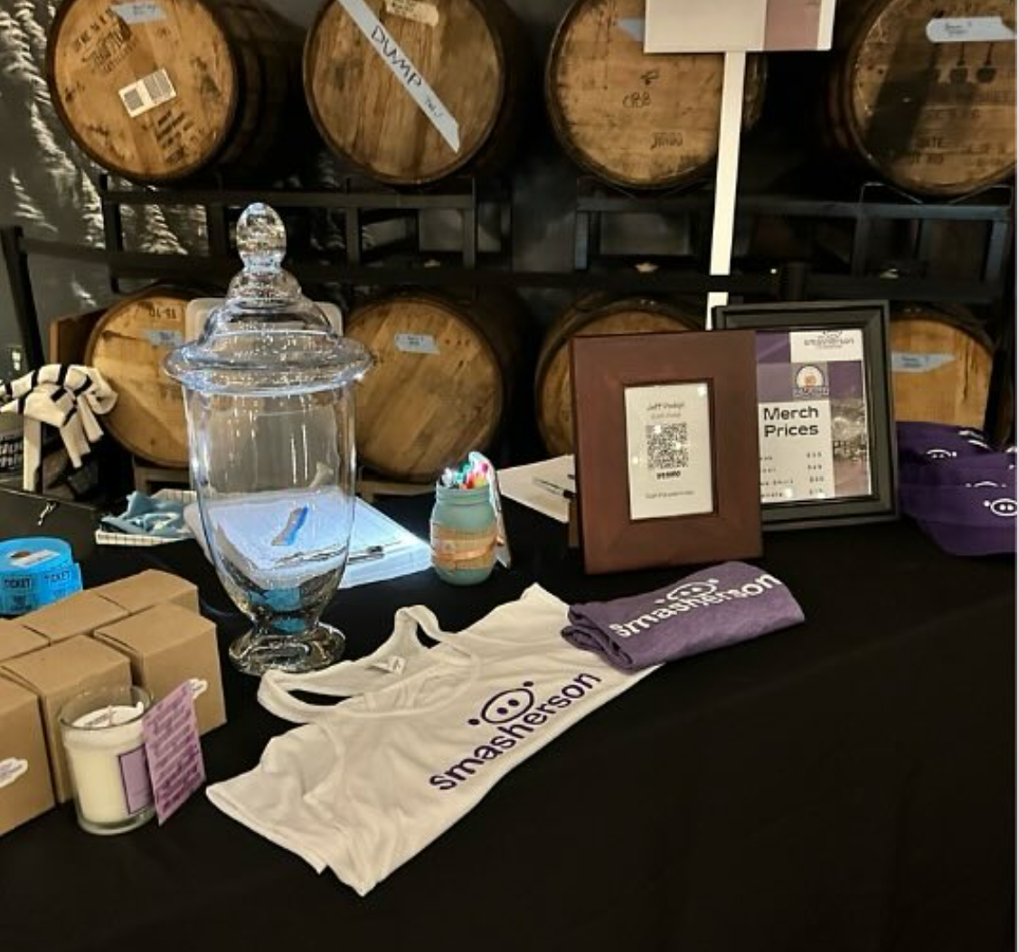 A table setup at an event booth with merchandise including a white tank top, purple wristbands, and small boxes, with a large glass jar and a small jar with candies on top. In the background, there are several wooden barrels and framed signs, one displaying 'Merch Prices'.