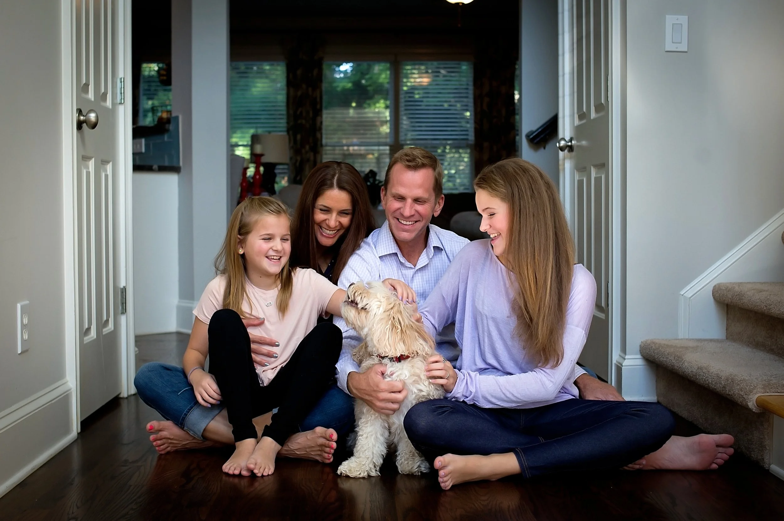 A family sits on the floor of their home, smiling and interacting with a small, fluffy dog. The family includes two women, a man, and a young girl, all appearing happy and engaged with the dog in a cozy living room with large windows and hardwood floors.