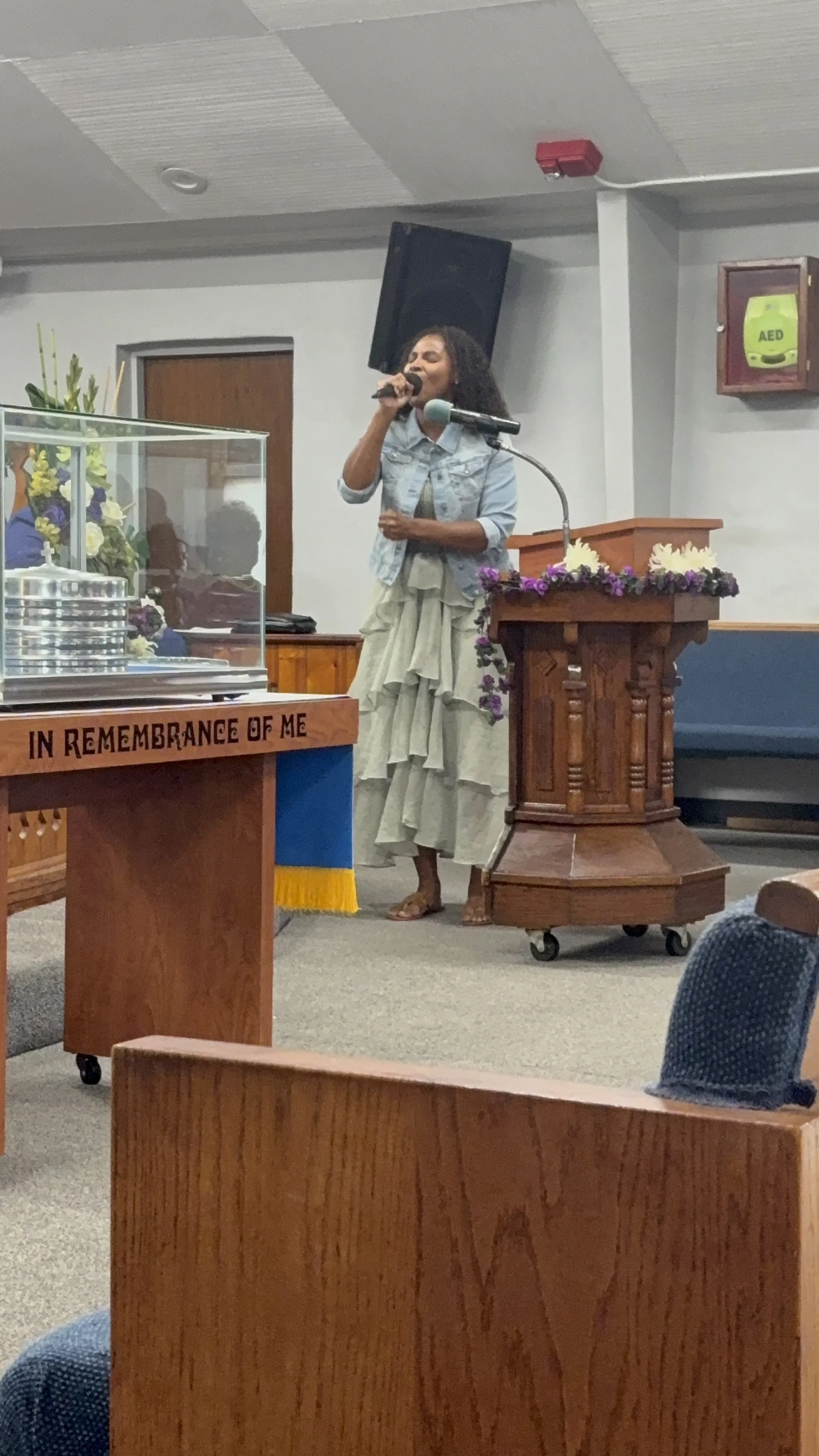 A woman singing into a microphone at a church or worship service. She is dressed in a denim jacket and a layered, ruffled skirt, standing next to a wooden podium decorated with purple and white flowers.