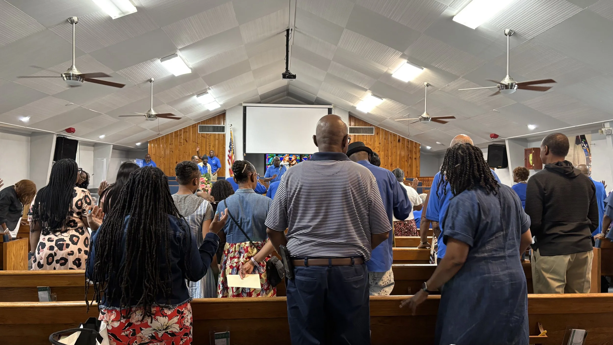 People standing in a church during a service, with some raising their hands in worship, facing the stage where a choir is performing.