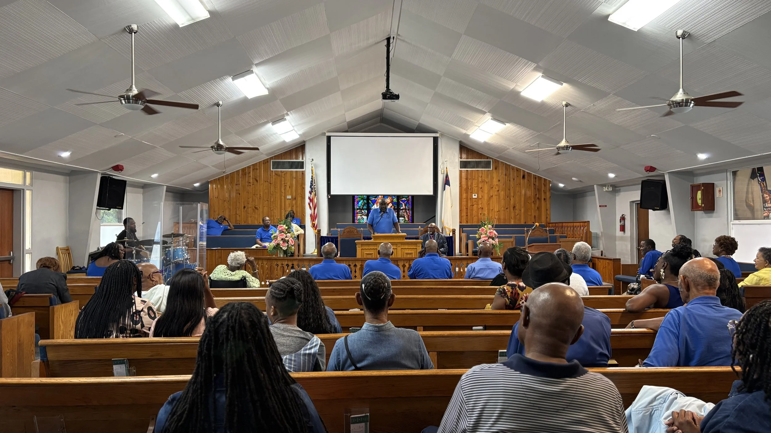 A church congregation seated on wooden pews, listening to a speaker at the pulpit. The church interior features a wooden background behind the pulpit, stained glass window, American flags, and floral arrangements.