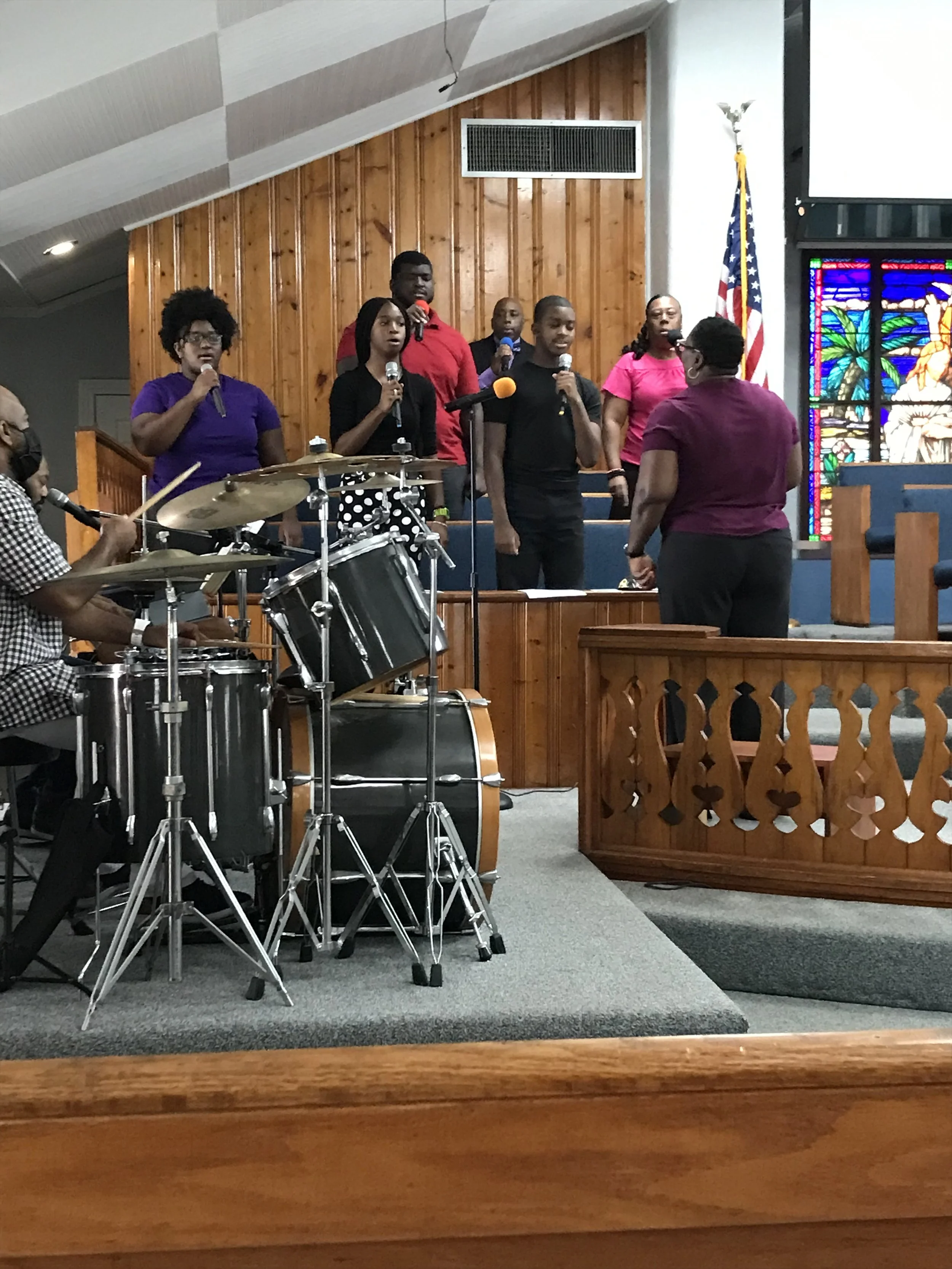 A choir of six singers standing on a church stage behind a conductor, with a drummer playing drums to the left, in front of stained glass windows and an American flag.