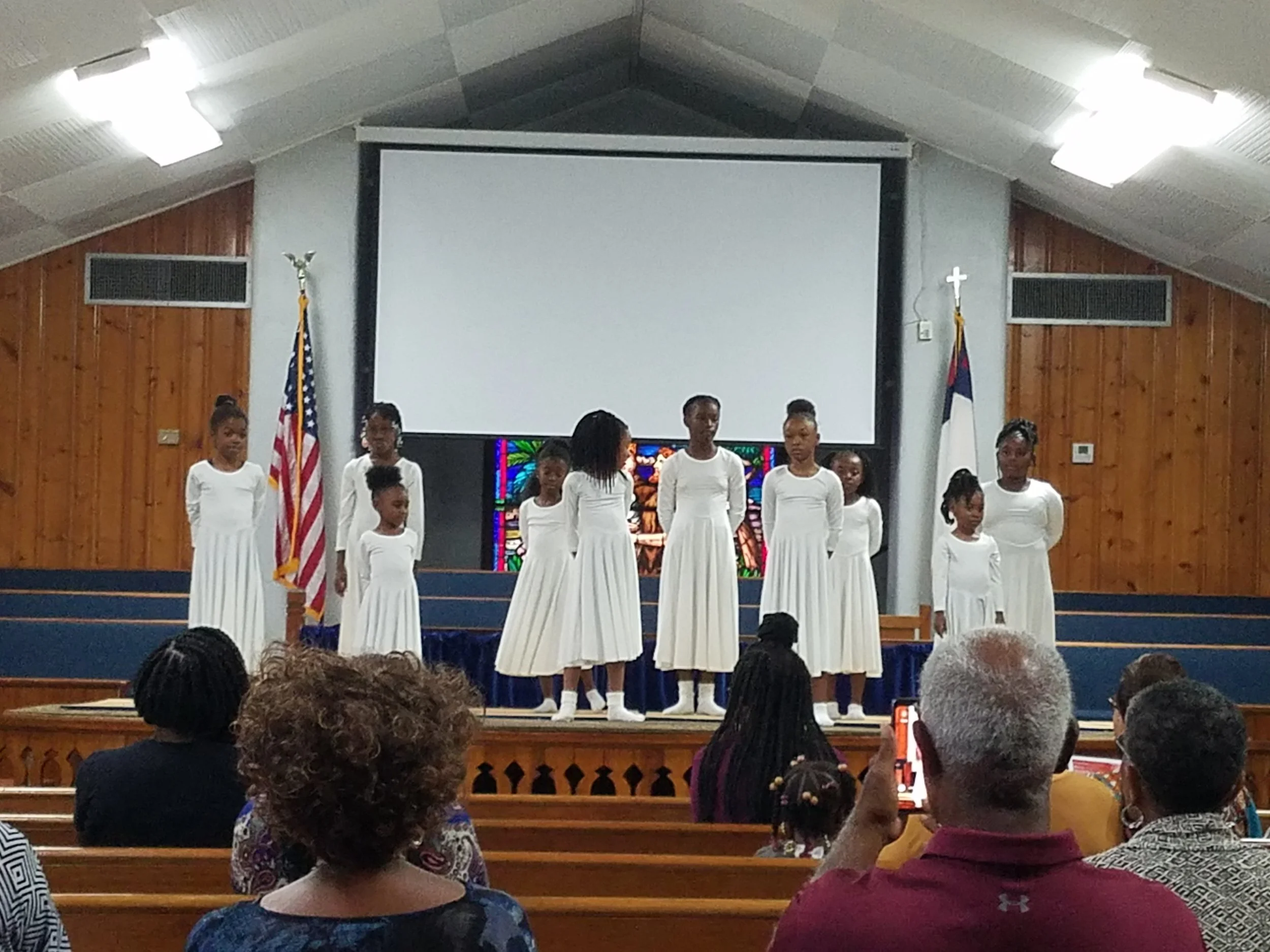 Young girls dressed in white robes performing on stage inside a church. Two flags, the American flag and the Christian flag, are positioned on either side of the stage. Audience members seated in the pews watching the performance.
