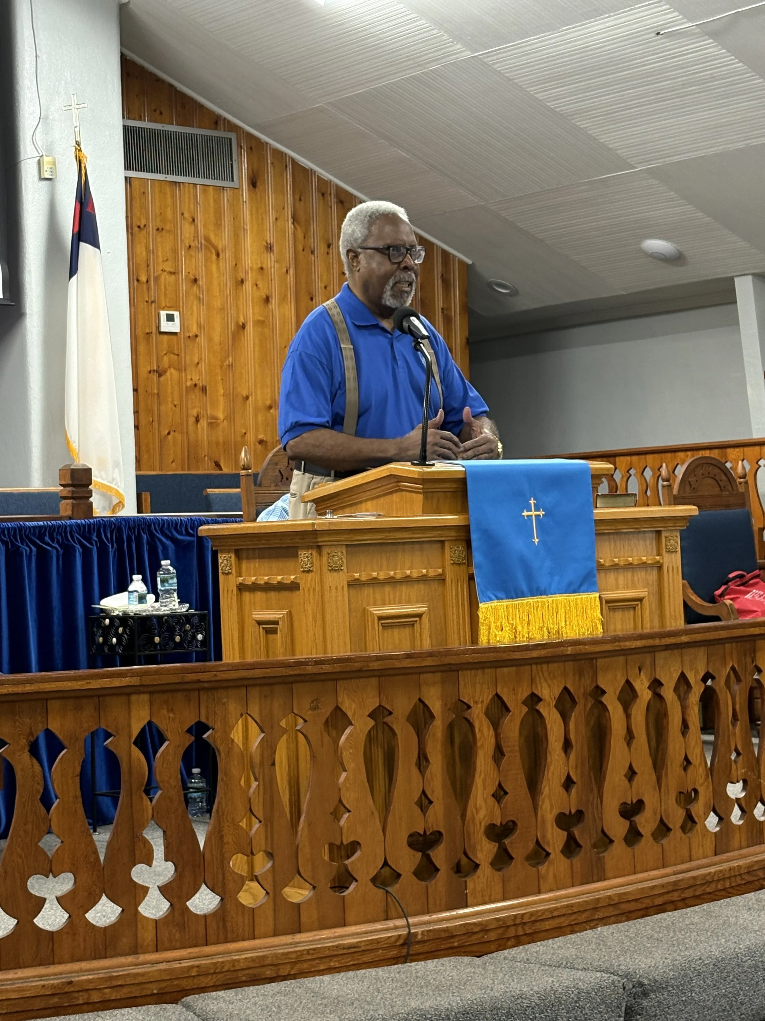 A man with gray hair, glasses, and a beard, dressed in a blue shirt with suspenders, stands at a wooden podium speaking into a microphone in a church setting. The background features wood-paneled walls, a flag, and a religious banner.