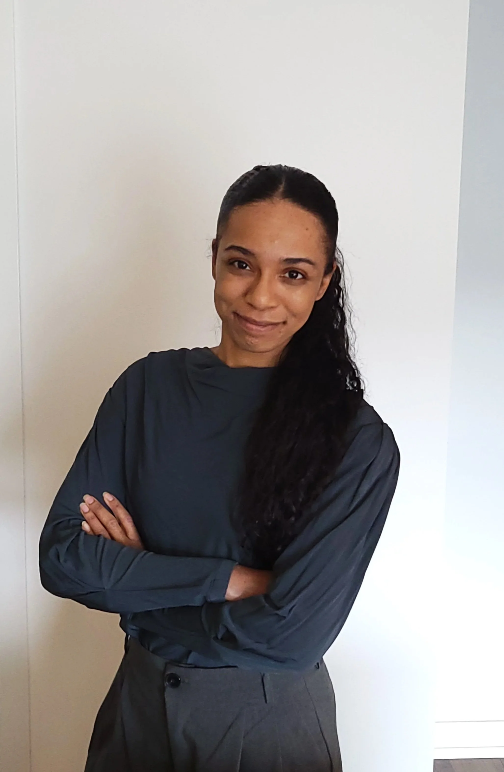 A woman with long, dark, curly hair, wearing a dark long-sleeve shirt and gray pants, standing with arms crossed, smiling at the camera in front of a plain white wall.