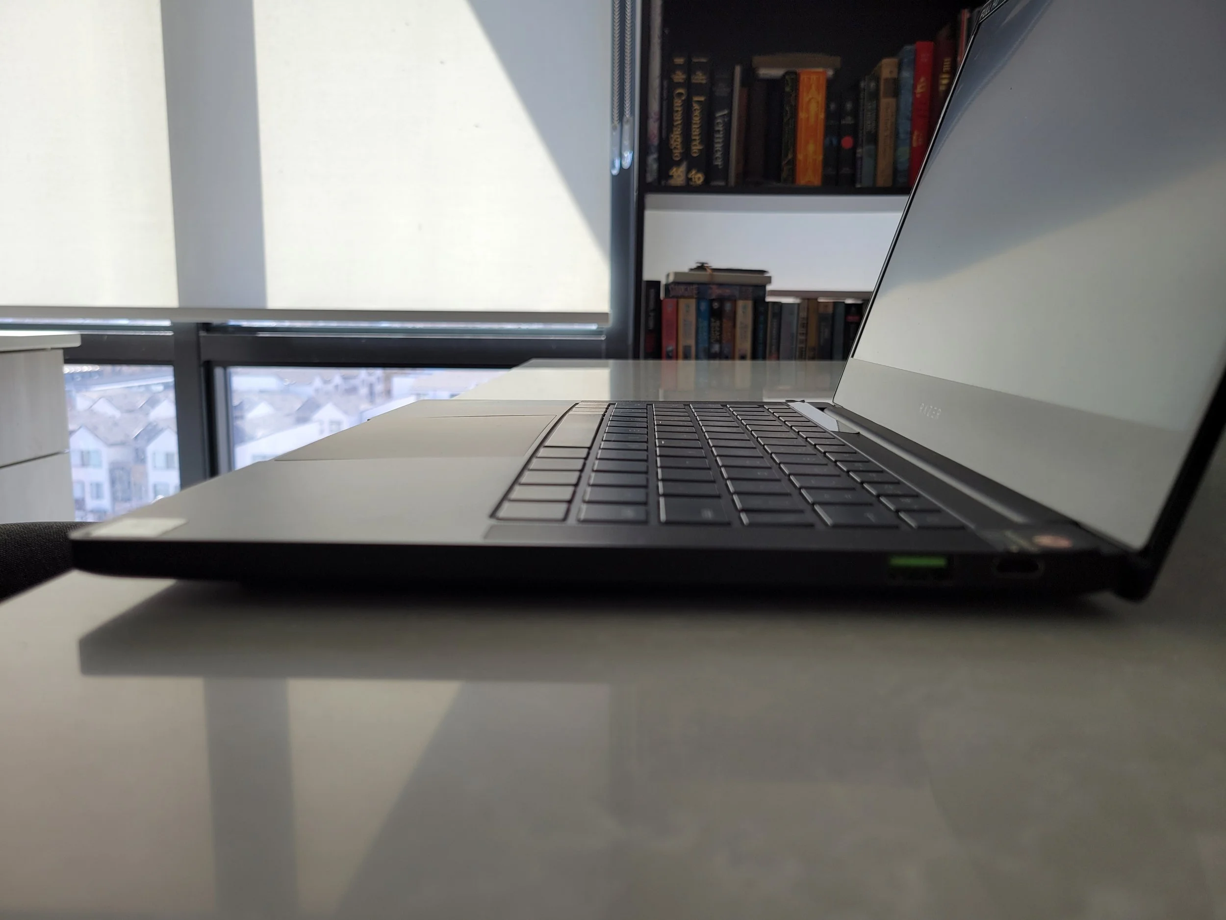 Side view of a modern laptop on a white desk in an office, with bookshelves and windows in the background.