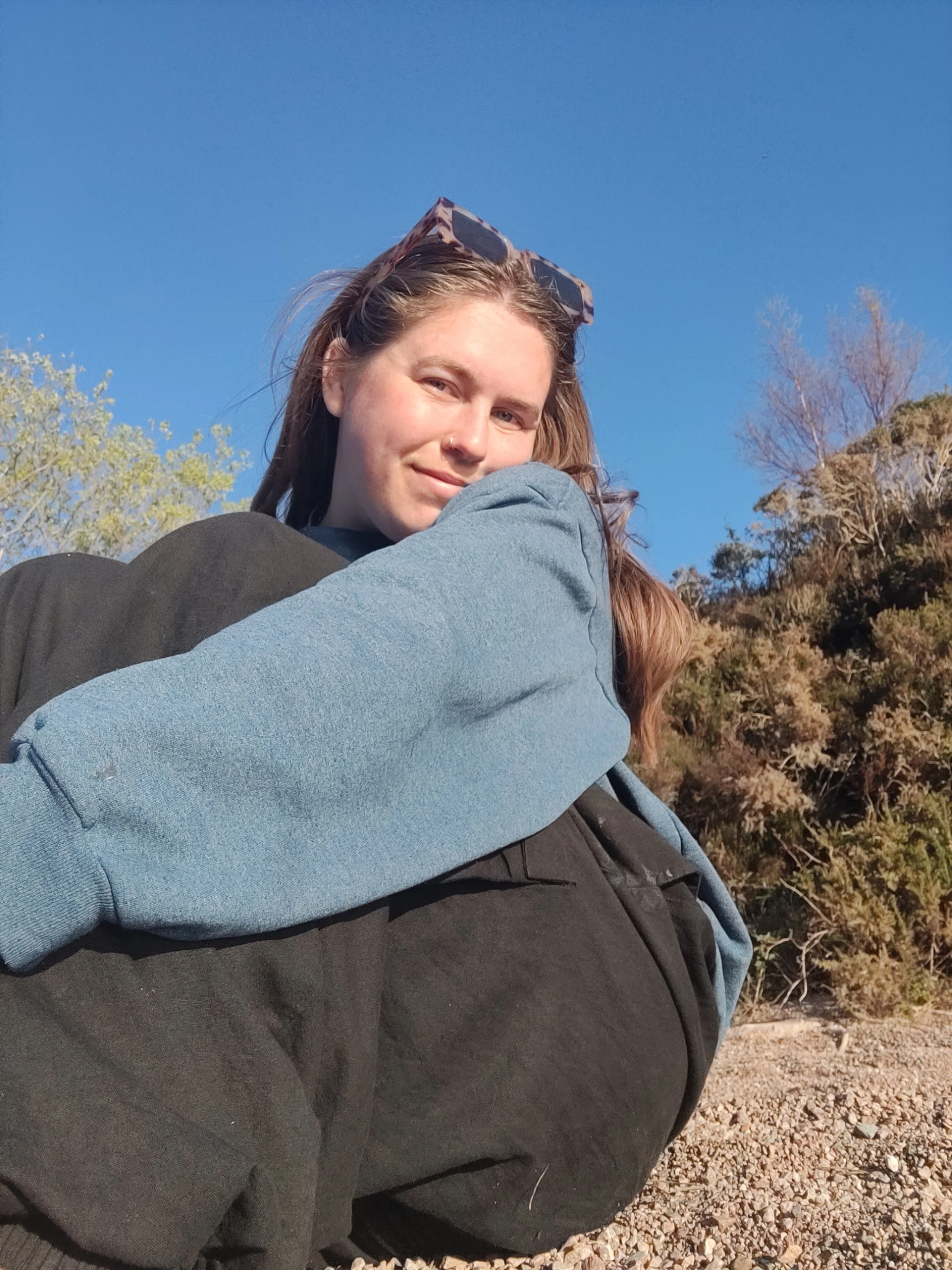 A woman with light brown hair and sunglasses on her head, wearing a black jacket and blue sweatshirt, is smiling while sitting outdoors on rocky terrain with bushes and a clear blue sky in the background.