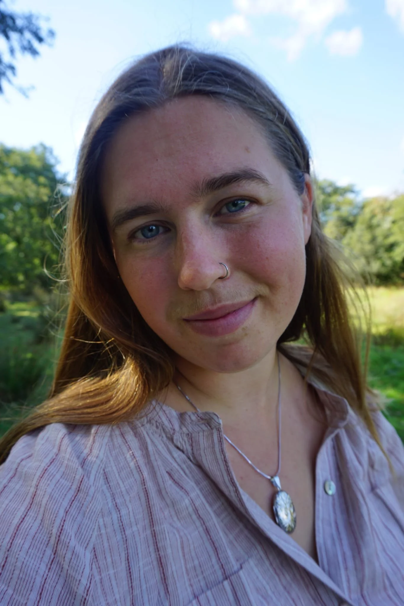 Close-up of a woman with long brown hair, blue eyes, and a nose ring, standing outdoors on a sunny day with trees and a clear sky in the background.