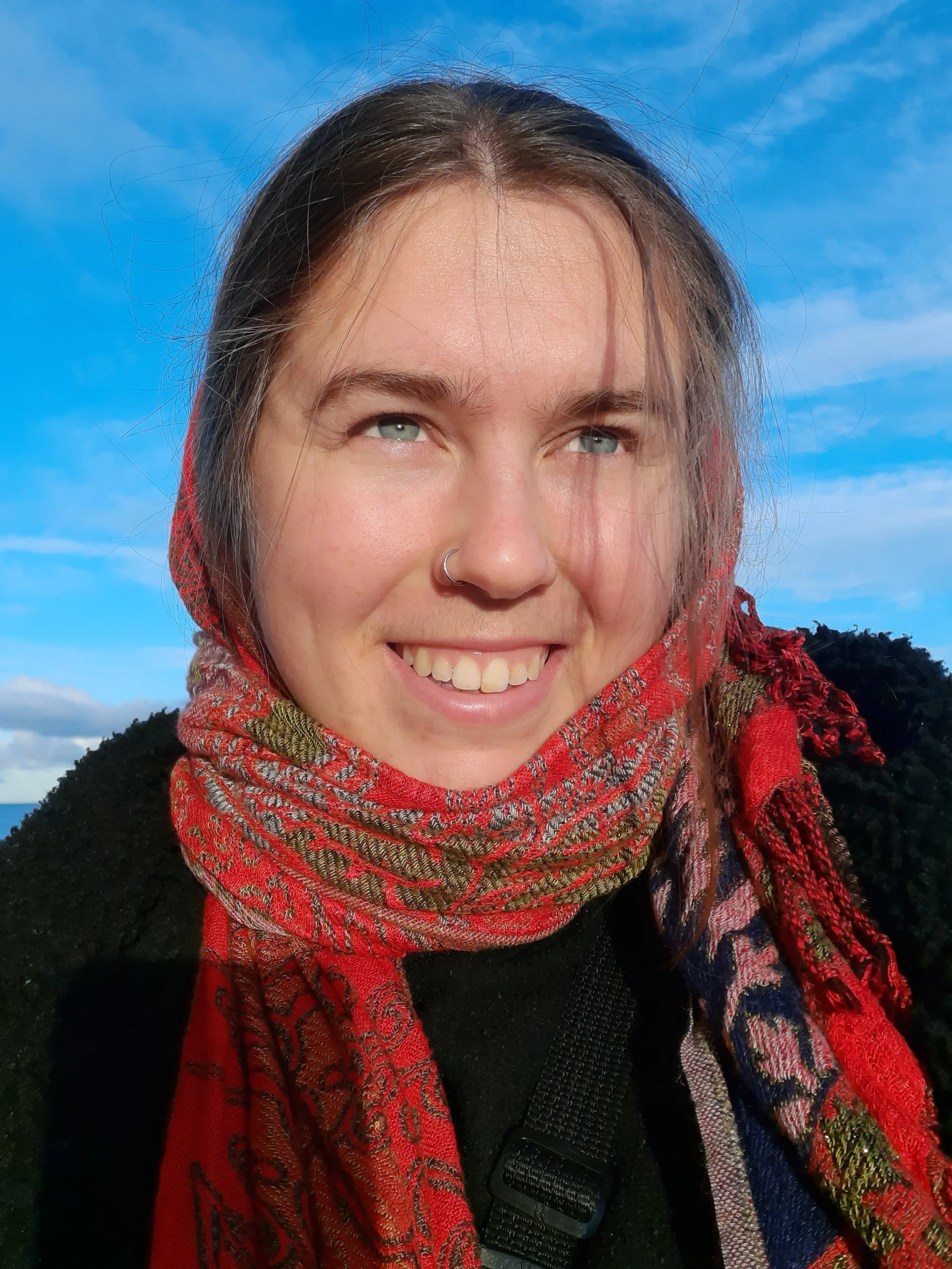 A young woman smiling outdoors with a blue sky background, wearing a colorful patterned scarf and a black top.