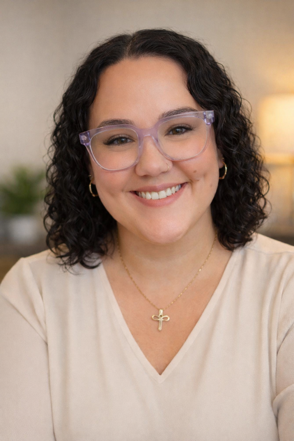 Smile of a young woman with curly dark hair, wearing clear glasses, a small nose ring, gold hoop earrings, a gold cross necklace, and a light-colored top, in an indoor setting with warm lighting.