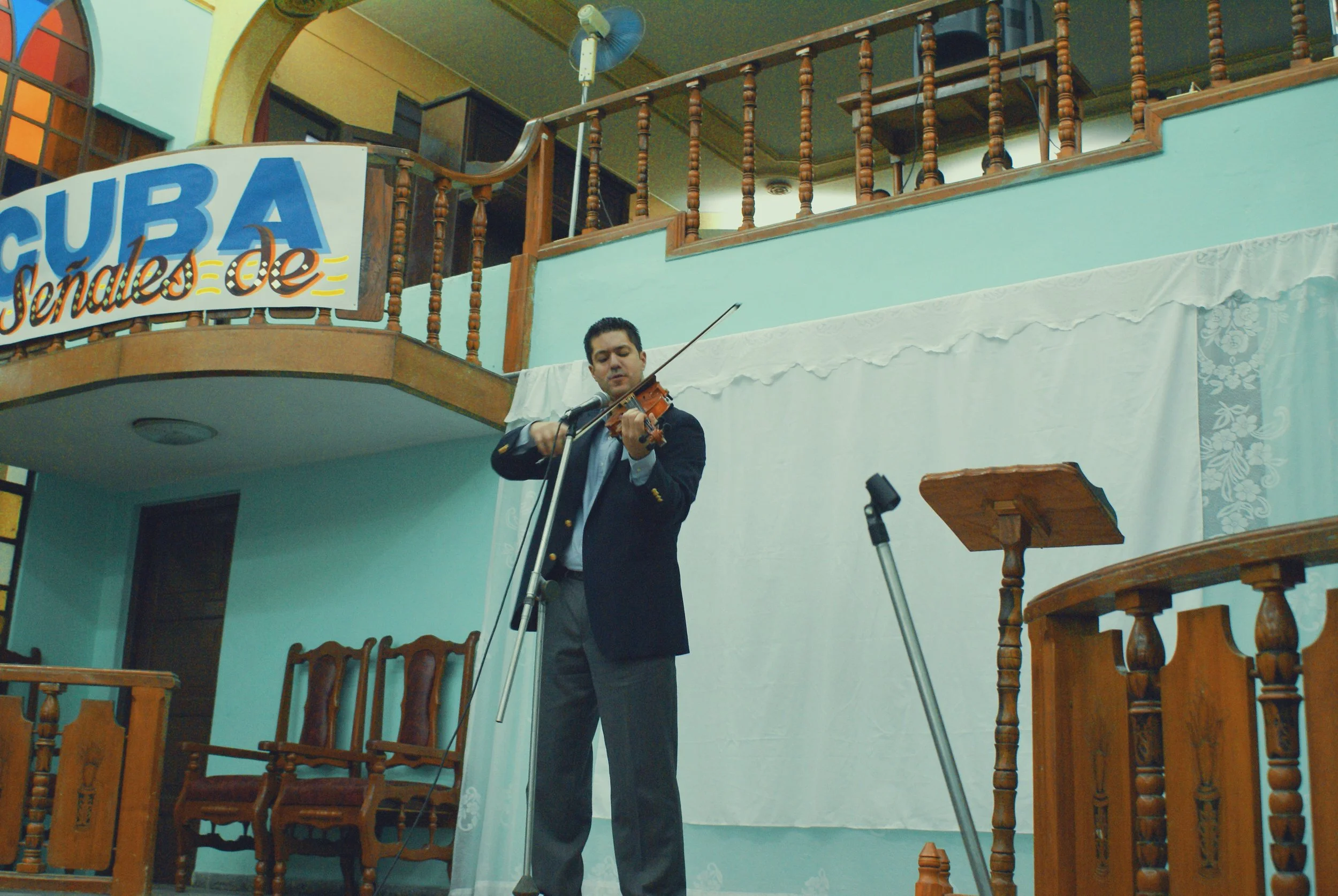 A man wearing a black blazer and gray pants playing a violin on a stage, with a microphone in front of him, in a room with wooden chairs and a balcony. A banner with the word 'CUBA' can be seen behind him.