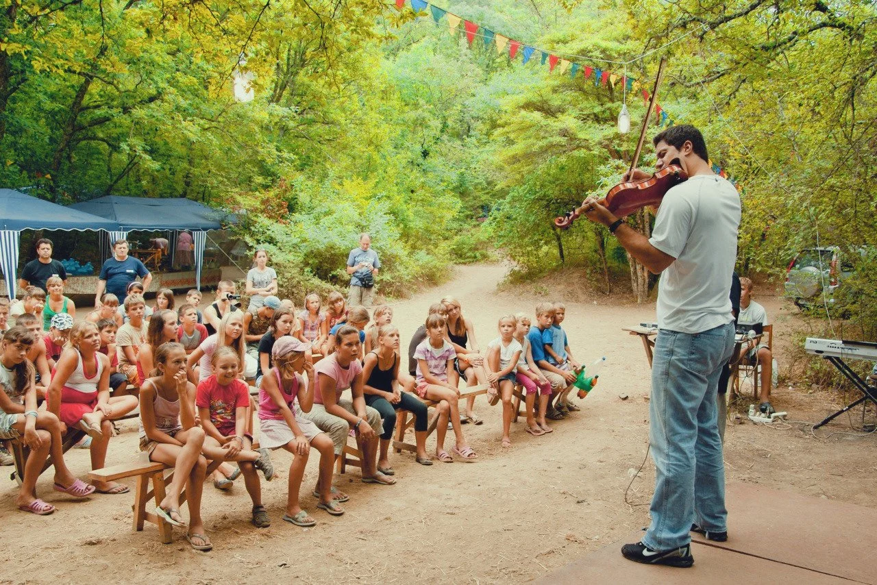 A man playing a violin on a stage outdoors while a group of children and a few adults sit on benches and watch, surrounded by trees and colorful flags.