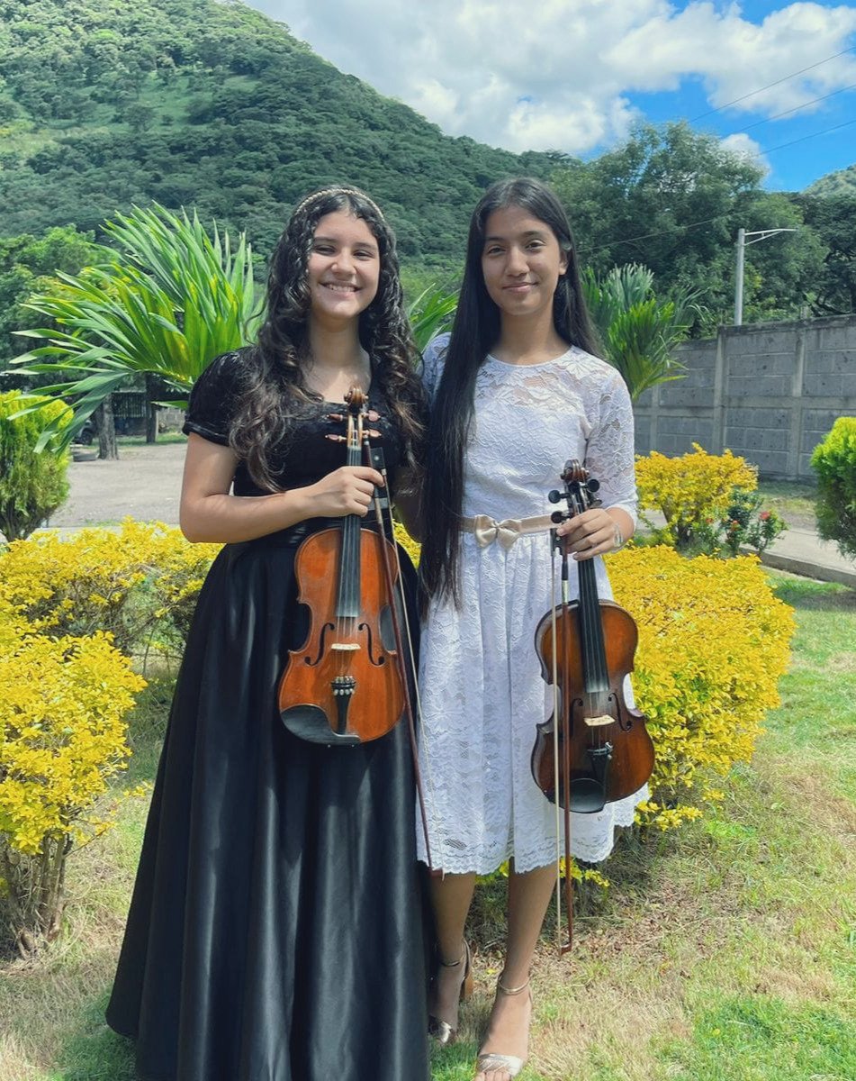 Two young women standing outdoors in a garden, holding violins, with mountains and a partly cloudy sky in the background.