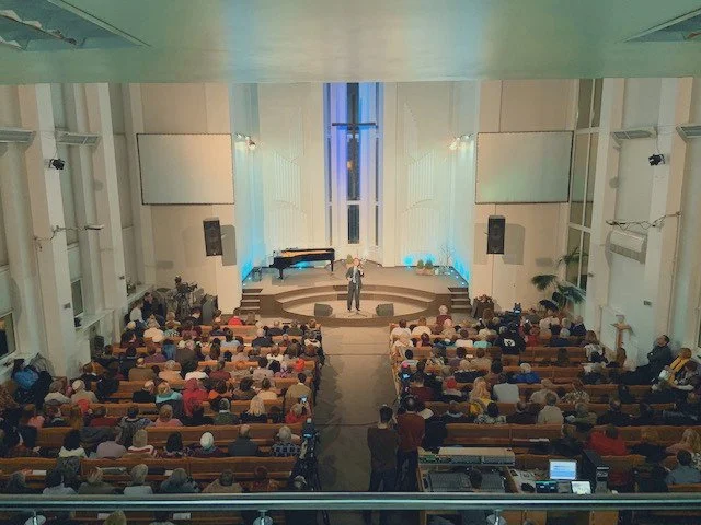 A speaker standing on a stage in a church or auditorium with a large audience seated in pews or chairs, and a piano on the stage.