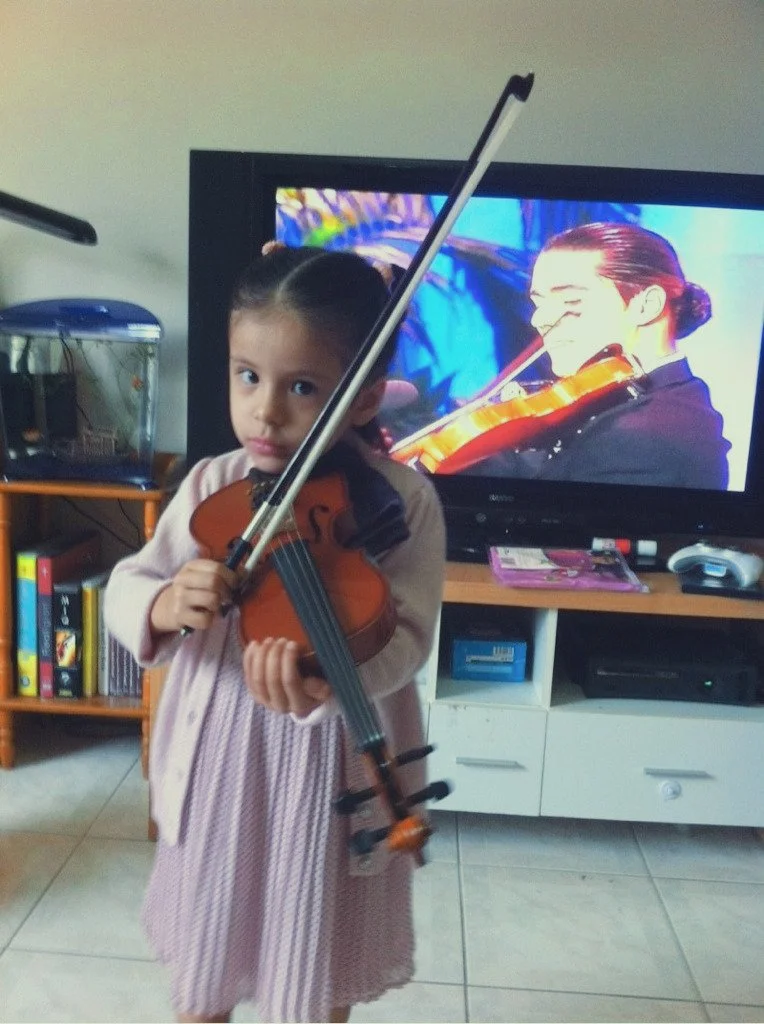 Young girl holding a violin and bow in a living room with a TV and bookshelf in the background.