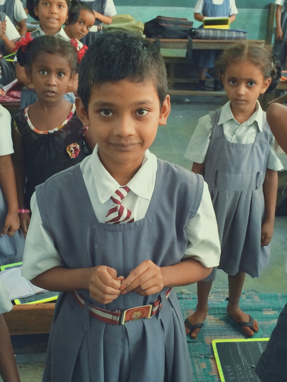 A group of young school children in uniform inside a classroom, with one girl in the foreground looking at the camera, smiling slightly, and wearing a gray dress with a white shirt and striped tie.