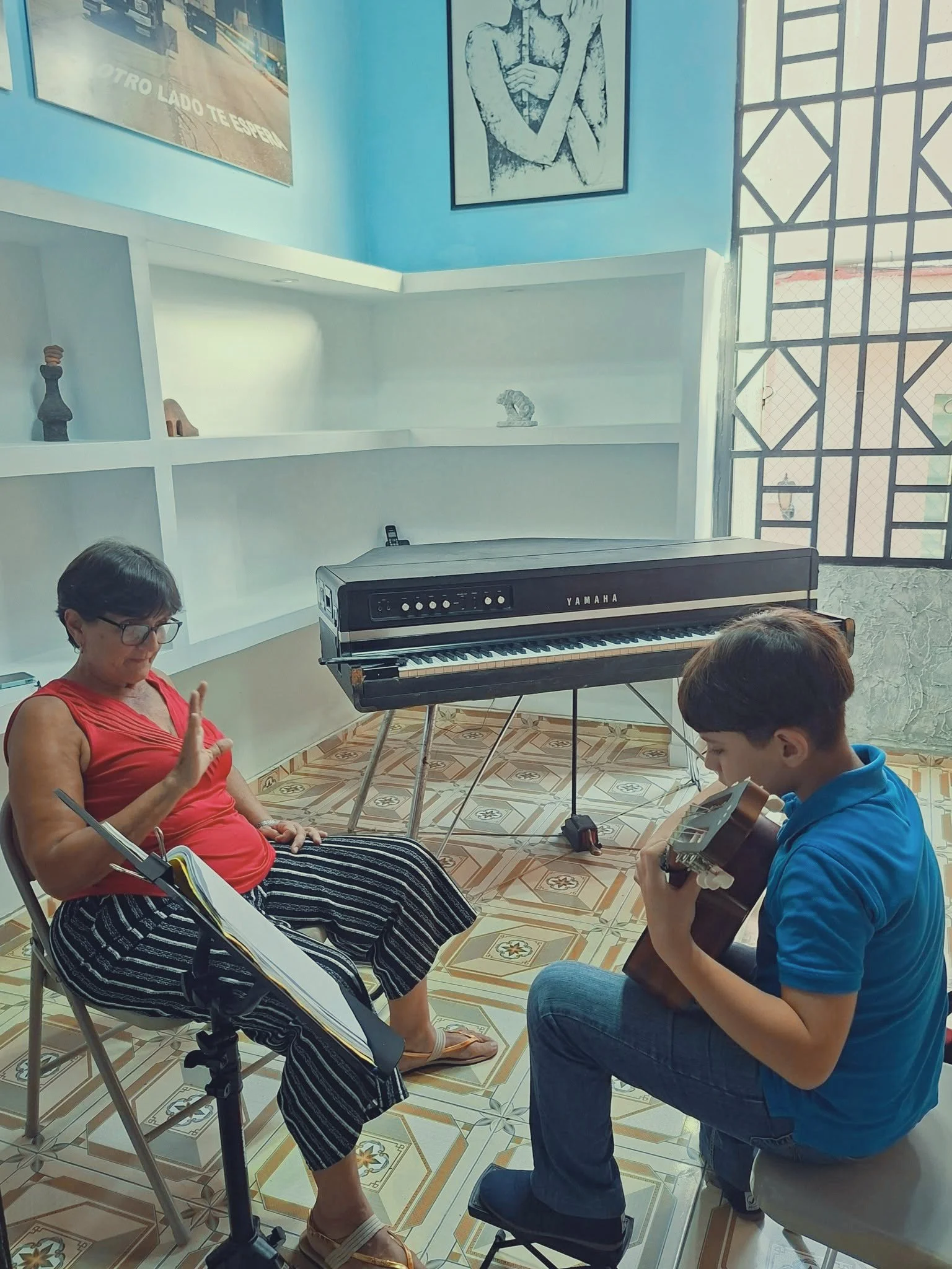 A woman with short hair sitting on a chair, playing a string instrument, while a young man in a blue shirt playing a guitar. There is a Yamaha keyboard beside them, and a music stand with sheet music in the foreground. The room has tiled floors and light blue walls decorated with posters and artwork.